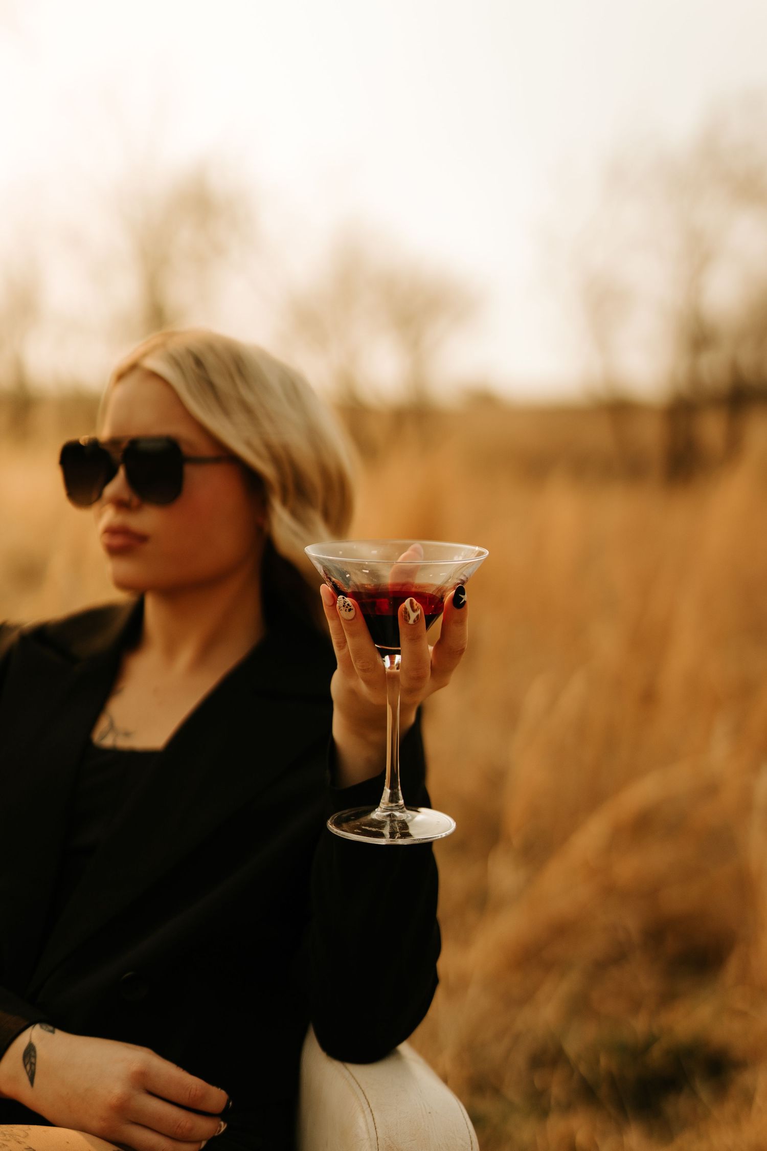 Blonde woman in sunglasses and black jacket holding a martini glass with red drink in a golden field.
