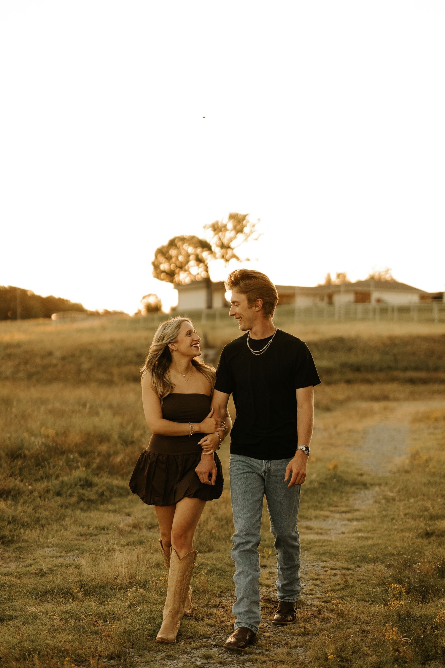 Couple walking hand-in-hand in a golden field at sunset, woman in black dress with cowboy boots, man in black tee and jeans.