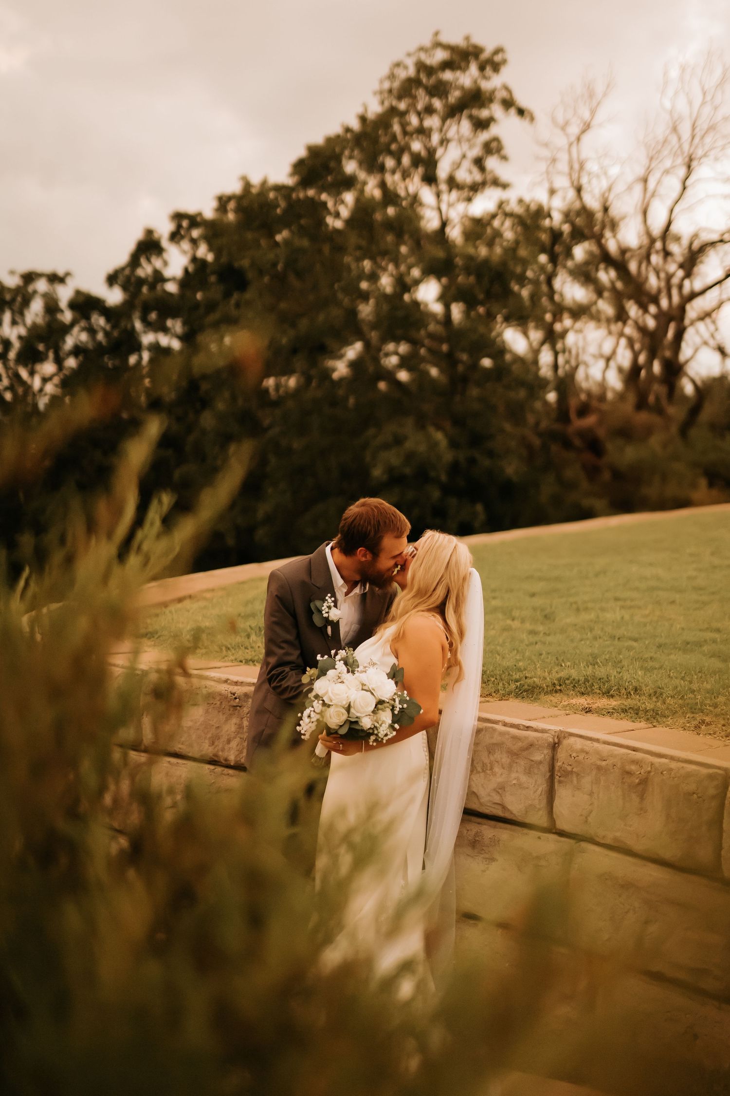Bride and groom sharing a kiss outdoors, bride holding white bouquet, stone wall and green trees in background.