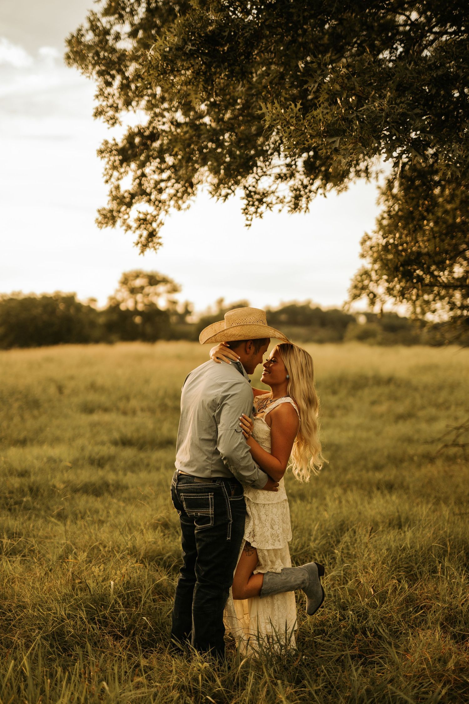 Couple sharing a romantic moment in a golden sunlit field, man in cowboy hat and woman in white dress embracing.