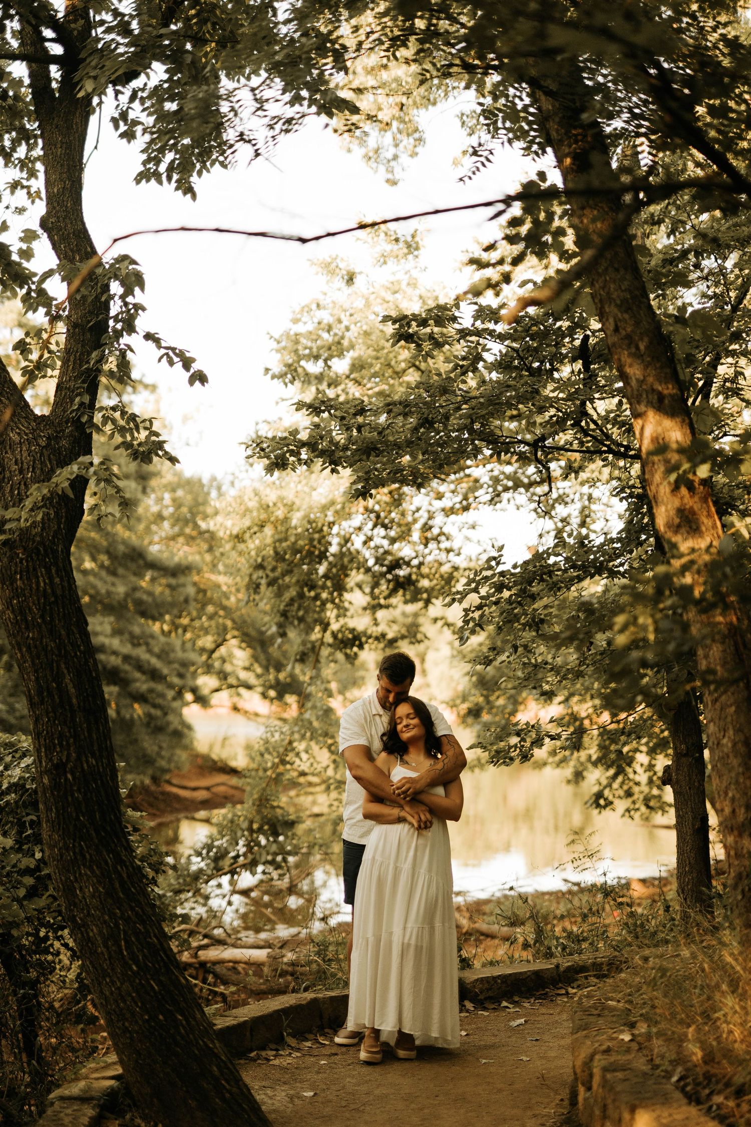 Couple embracing on a wooded path near a reflective pond, woman in white dress, warm golden light filtering through trees.