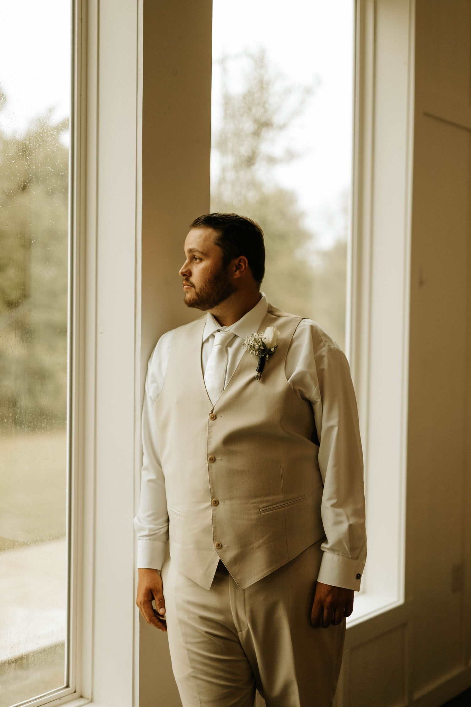 Groom in beige suit vest gazes through window in contemplative pose.