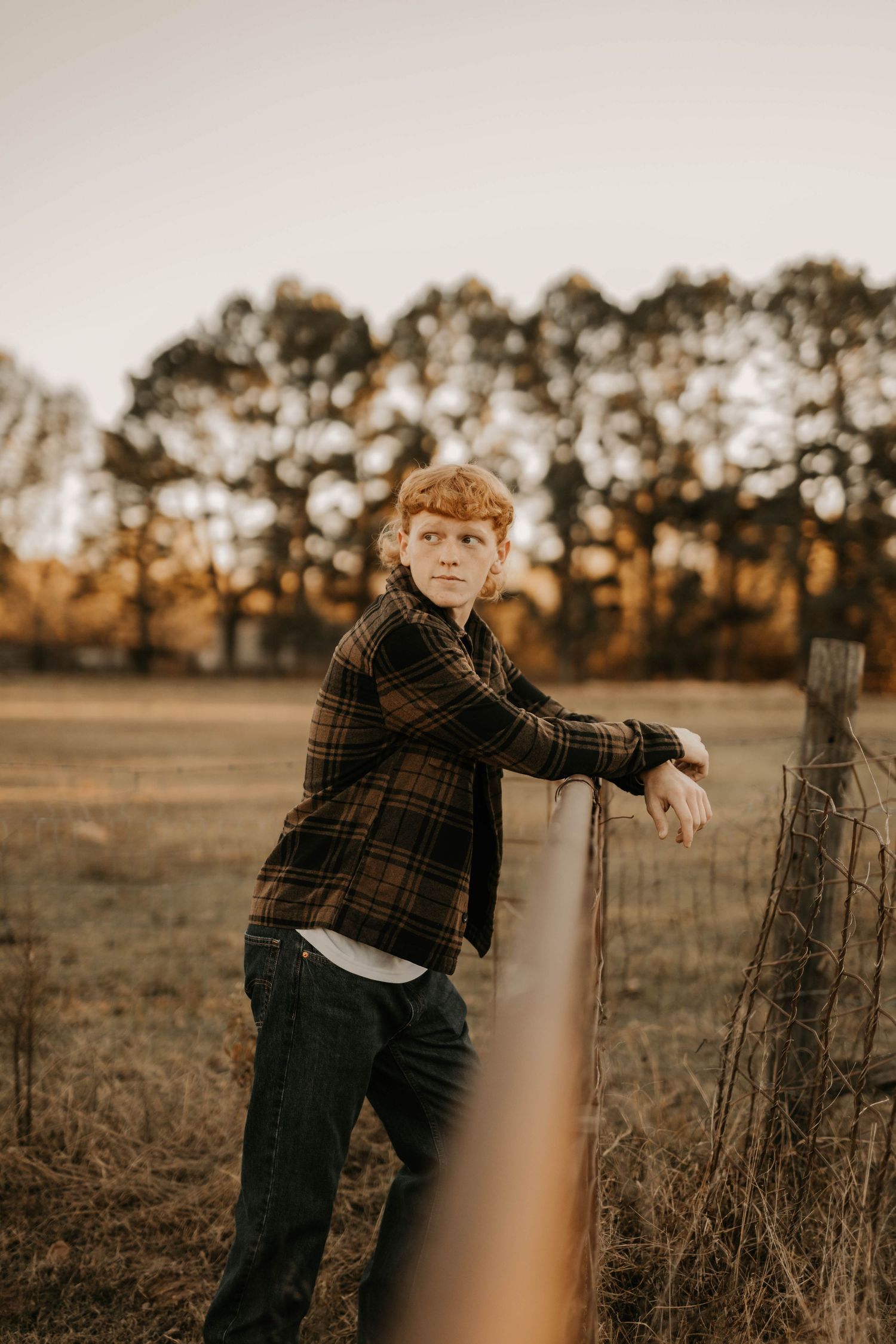 Person in plaid flannel leans against wooden fence post at sunset with trees in background.