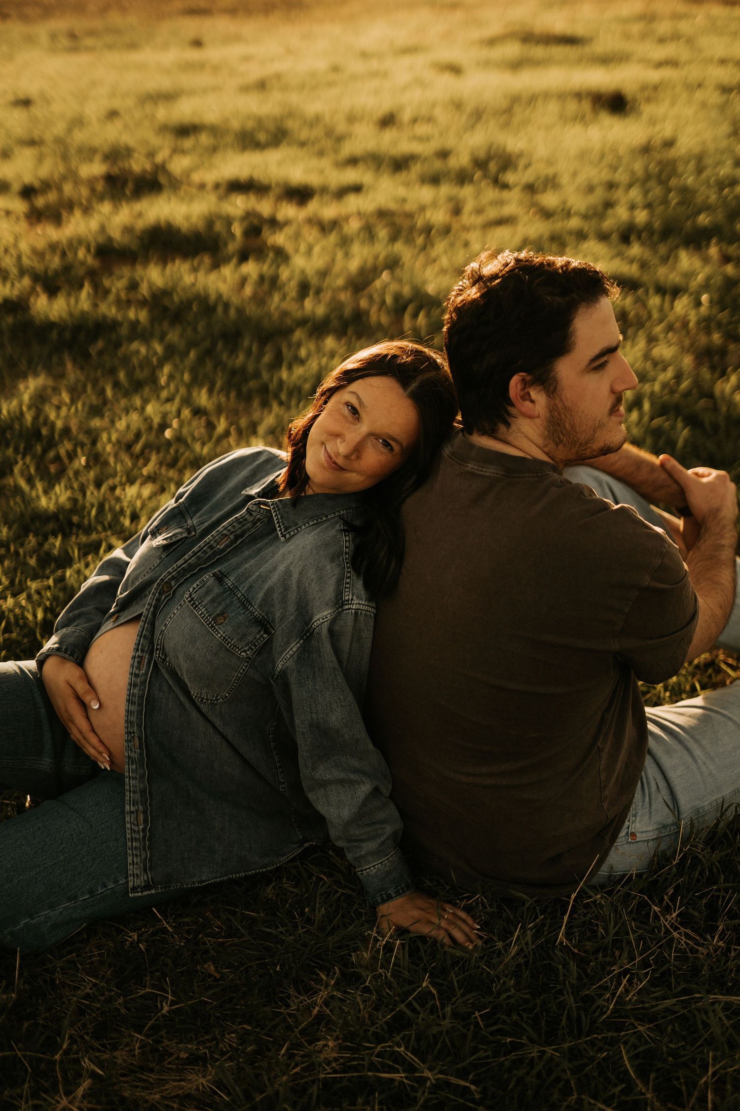 Pregnant woman in denim jacket leans on partner's back while sitting in golden grass during sunset maternity photoshoot.