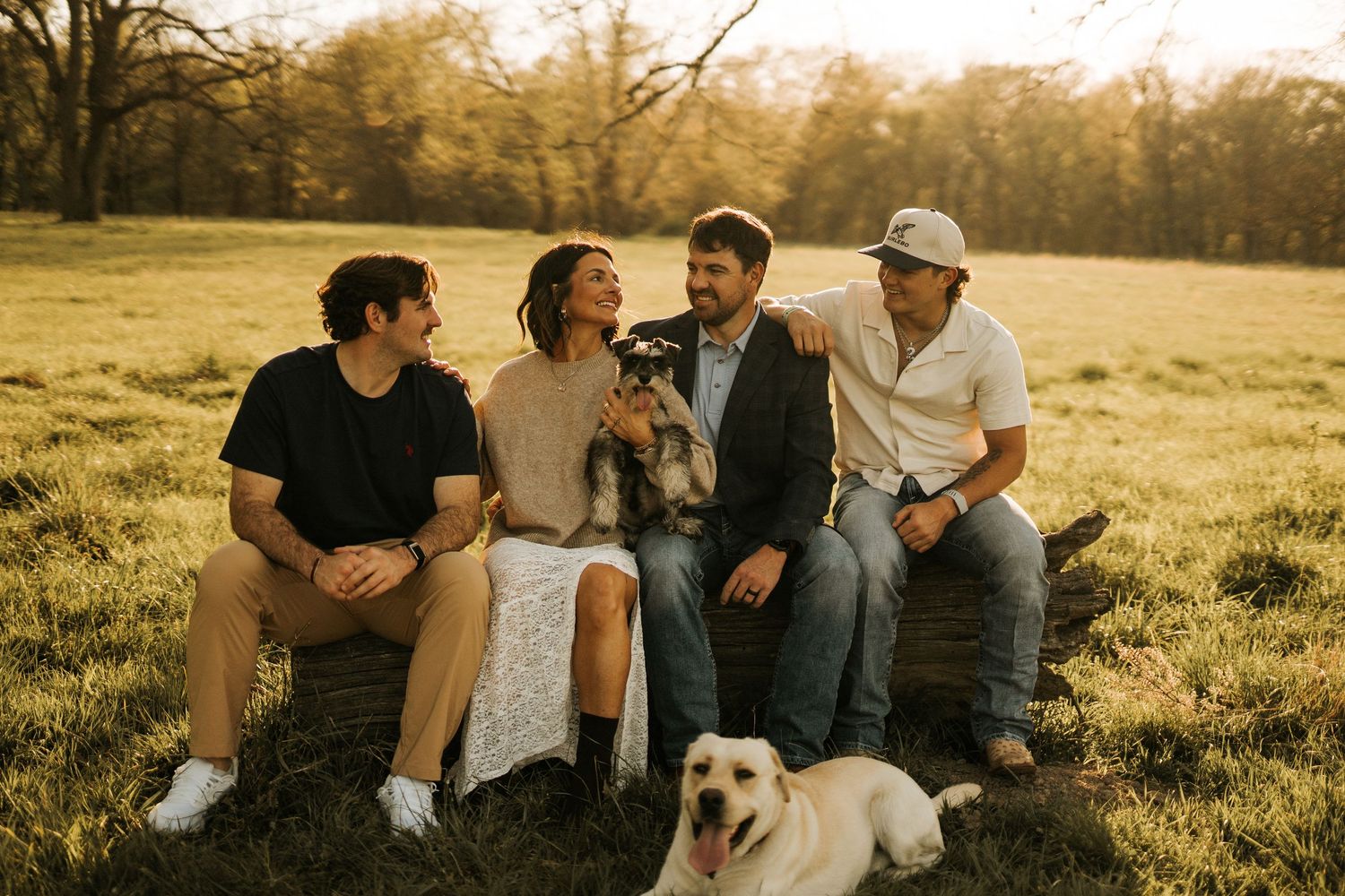 Family of four smiling together outdoors at golden hour with two dogs on a grassy field.