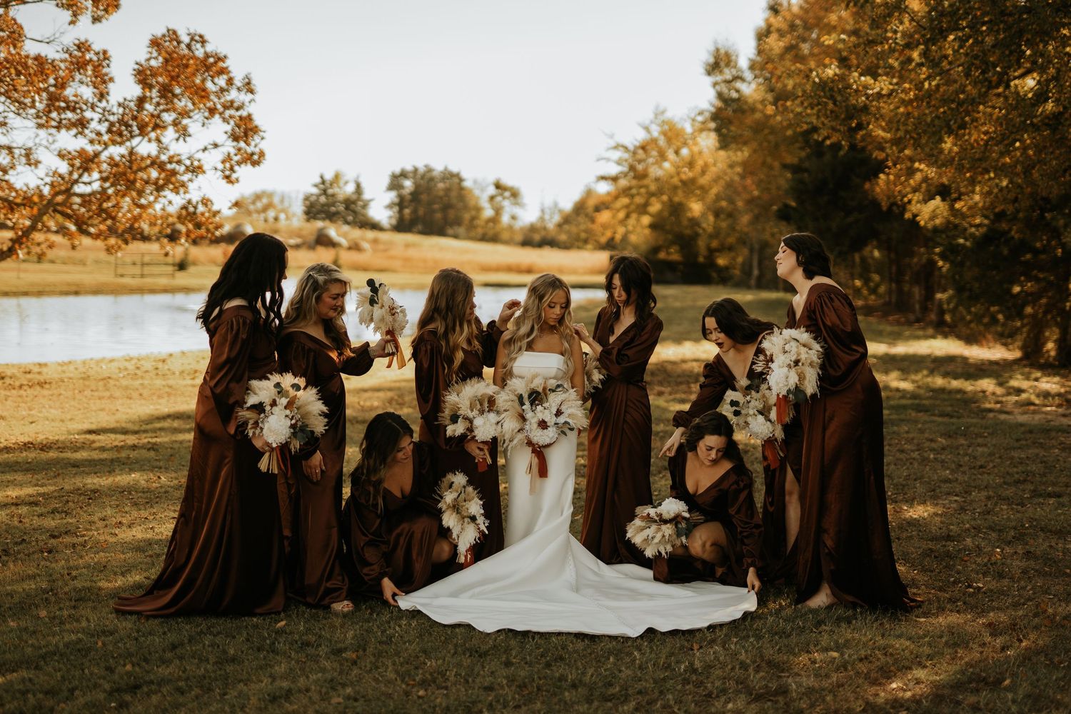 Bride in white gown surrounded by bridesmaids in brown dresses holding white bouquets outdoors in autumn setting.