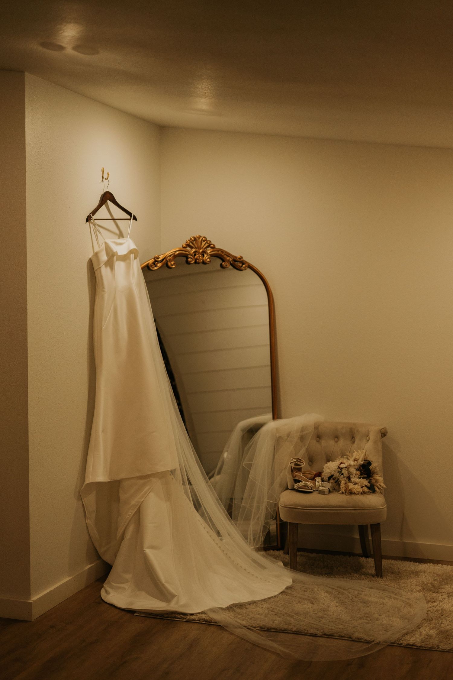 Elegant white wedding dress with long train hanging on a wooden hanger beside a gold-framed mirror in a warmly lit bridal suite.