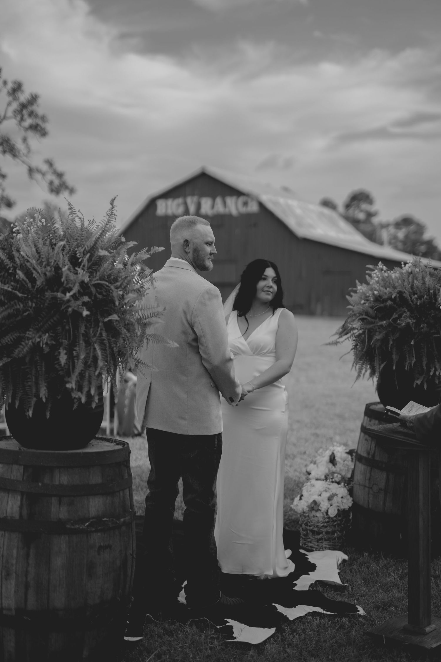 Bride and groom hold hands at outdoor barn wedding ceremony at Big V Ranch, surrounded by rustic barrels and lush greenery.