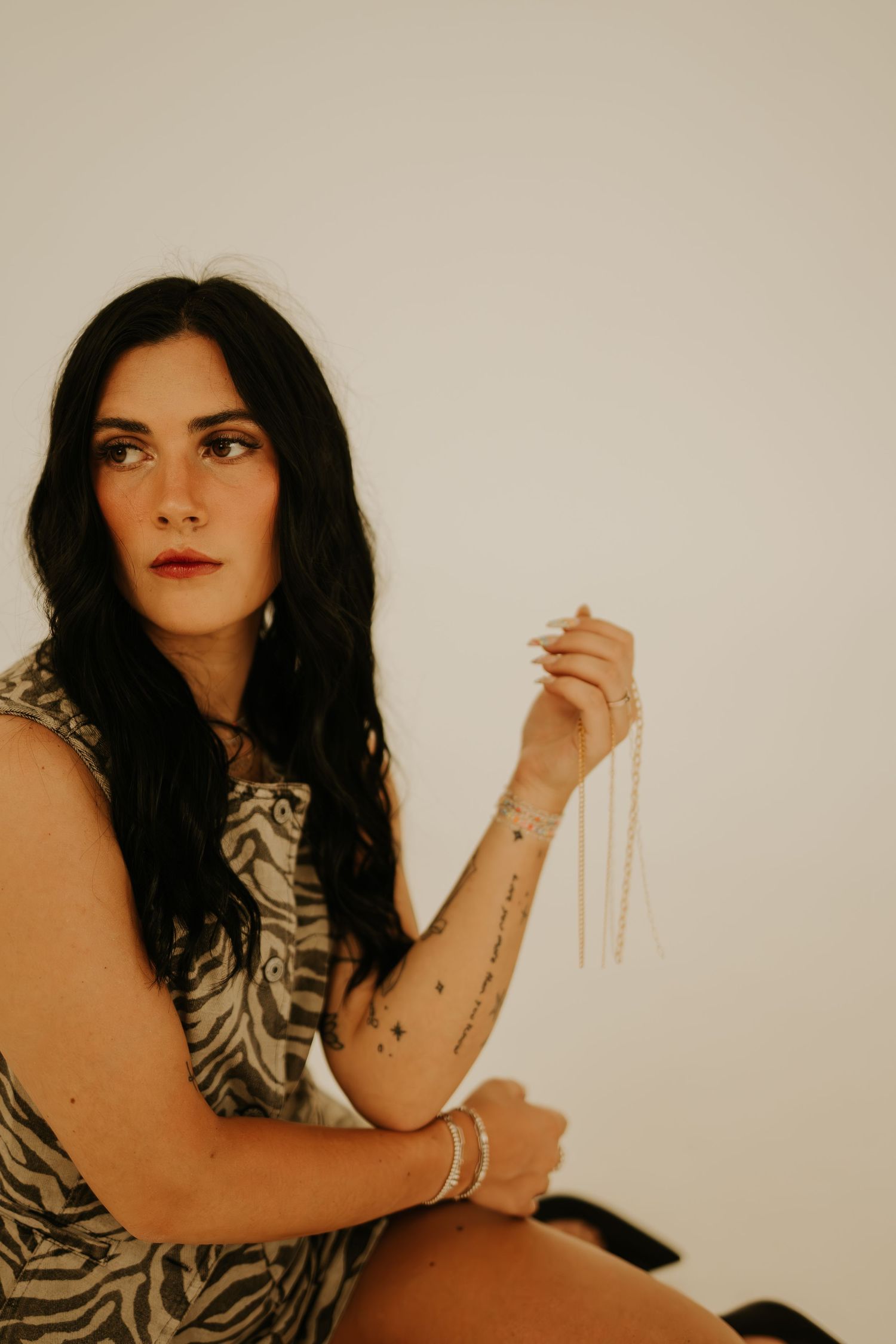 Dark-haired woman in zebra print dress holds pearl necklace, showing arm tattoos, seated against white background.