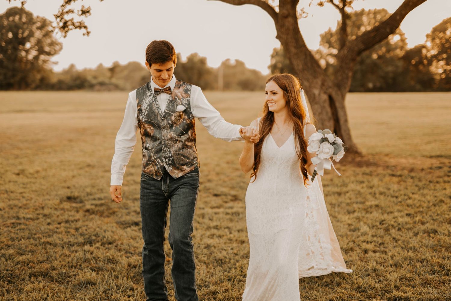 Couple walks together in rustic field with bride in white dress and groom wearing western-style vest.