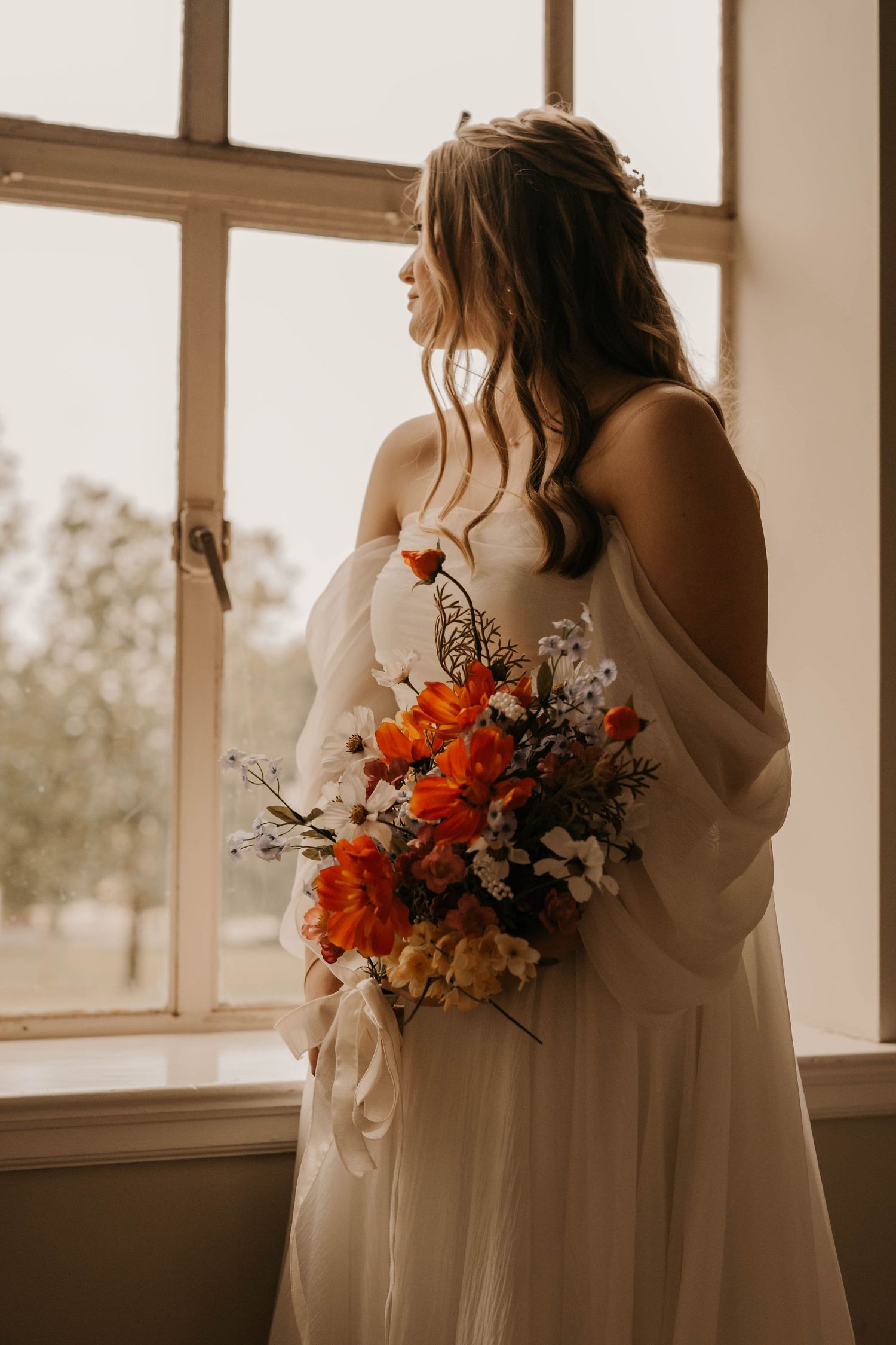 A bride in white holds vibrant orange and white flowers by a sunny window.