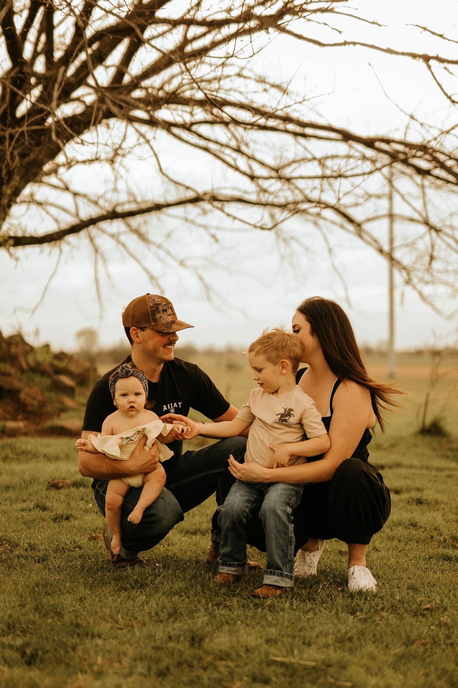 Family sits together on grass under bare tree branches during golden hour photo session.