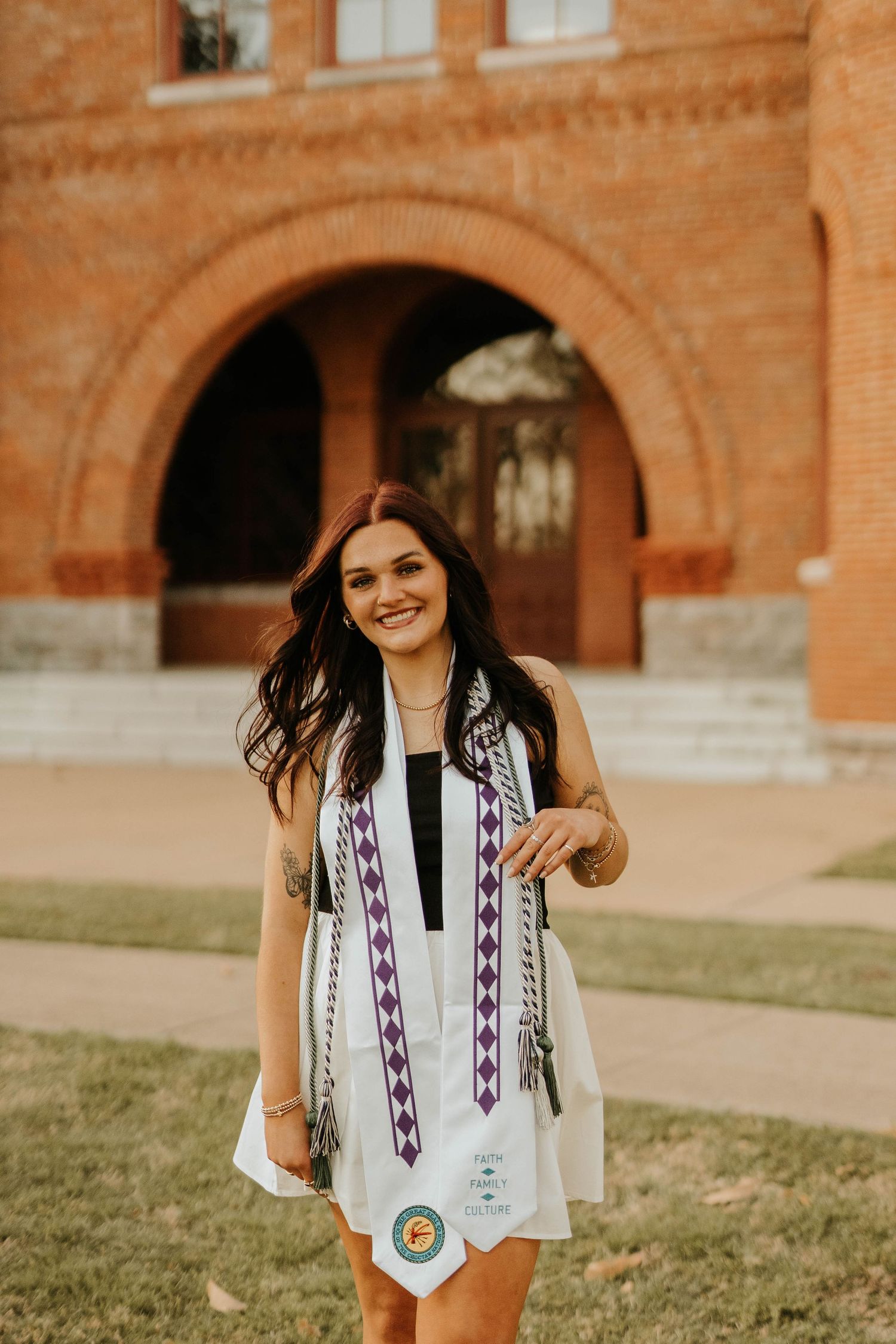 Graduate in summer dress and decorative stole smiles in front of historic brick building.