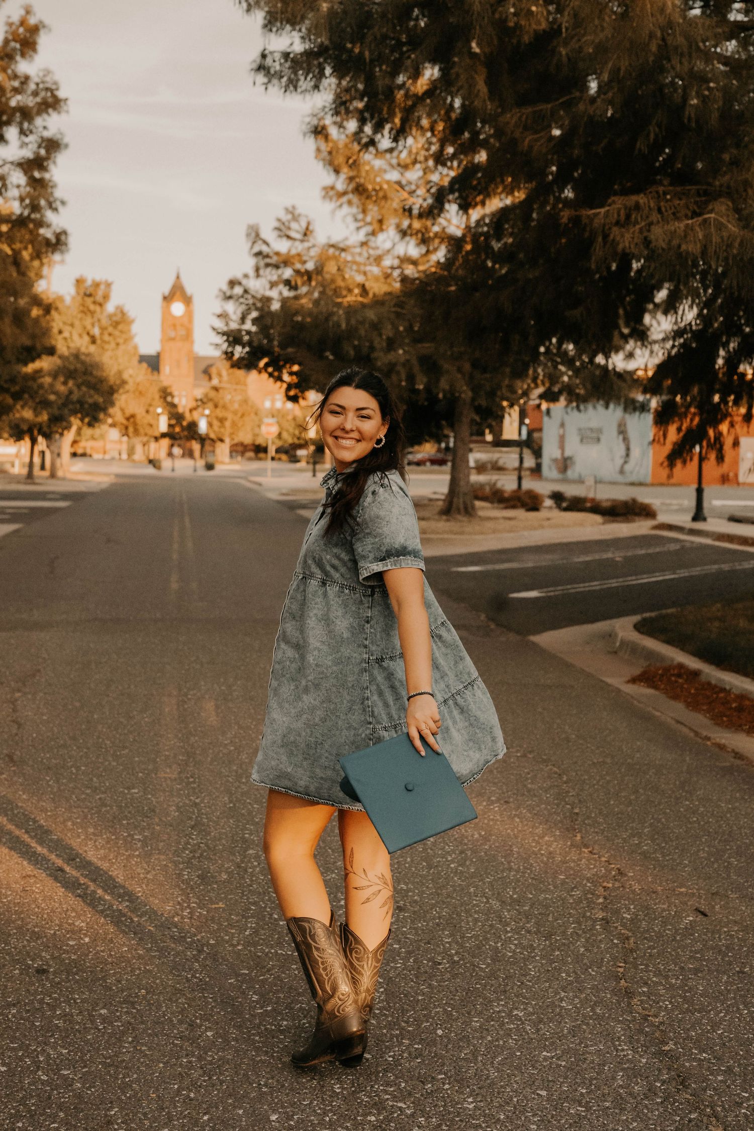 Person in denim dress twirls on street with graduation cap and historic clock tower in background.