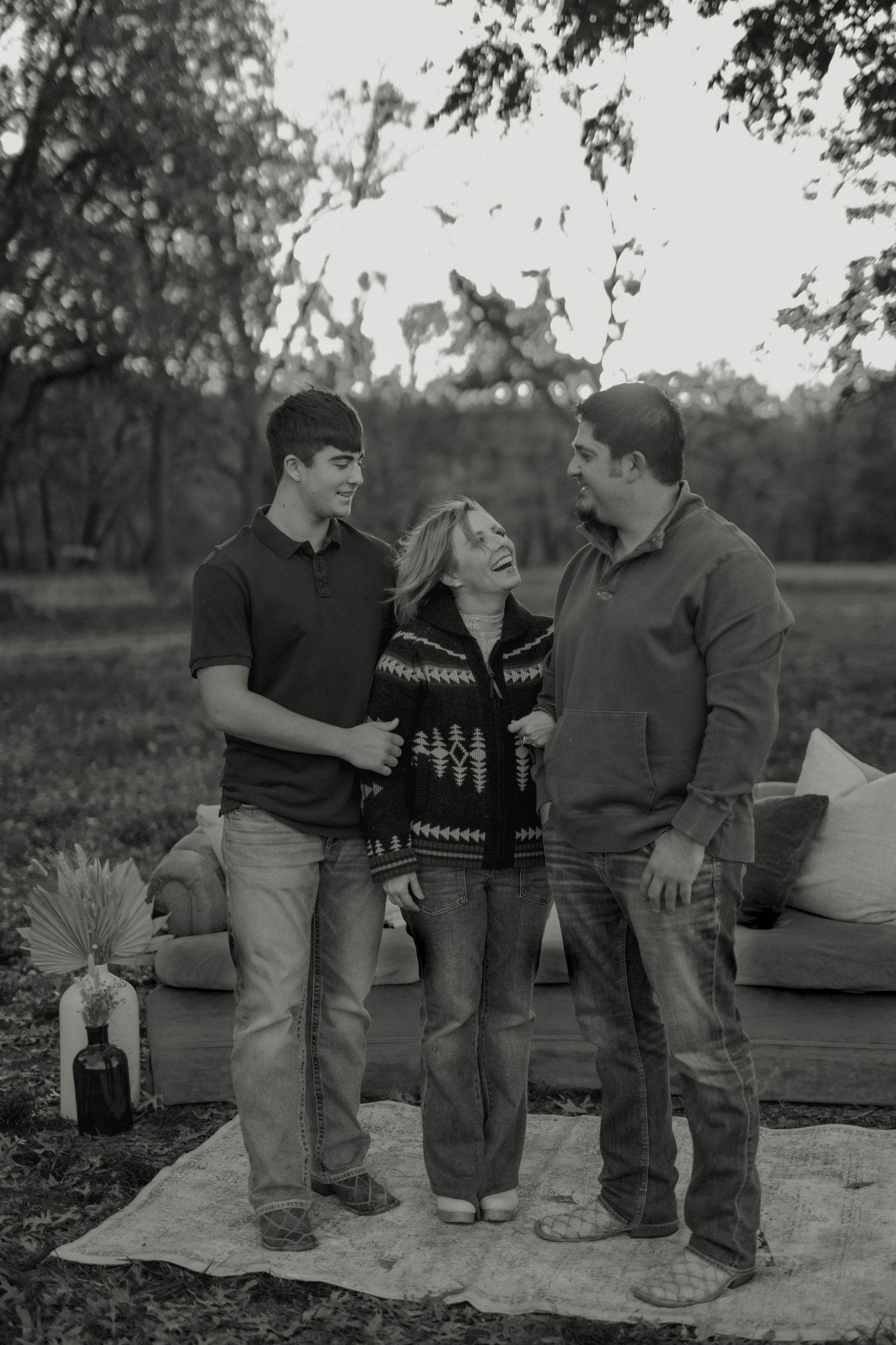 Black and white family portrait of three people laughing outdoors near a couch on a blanket in a park setting.