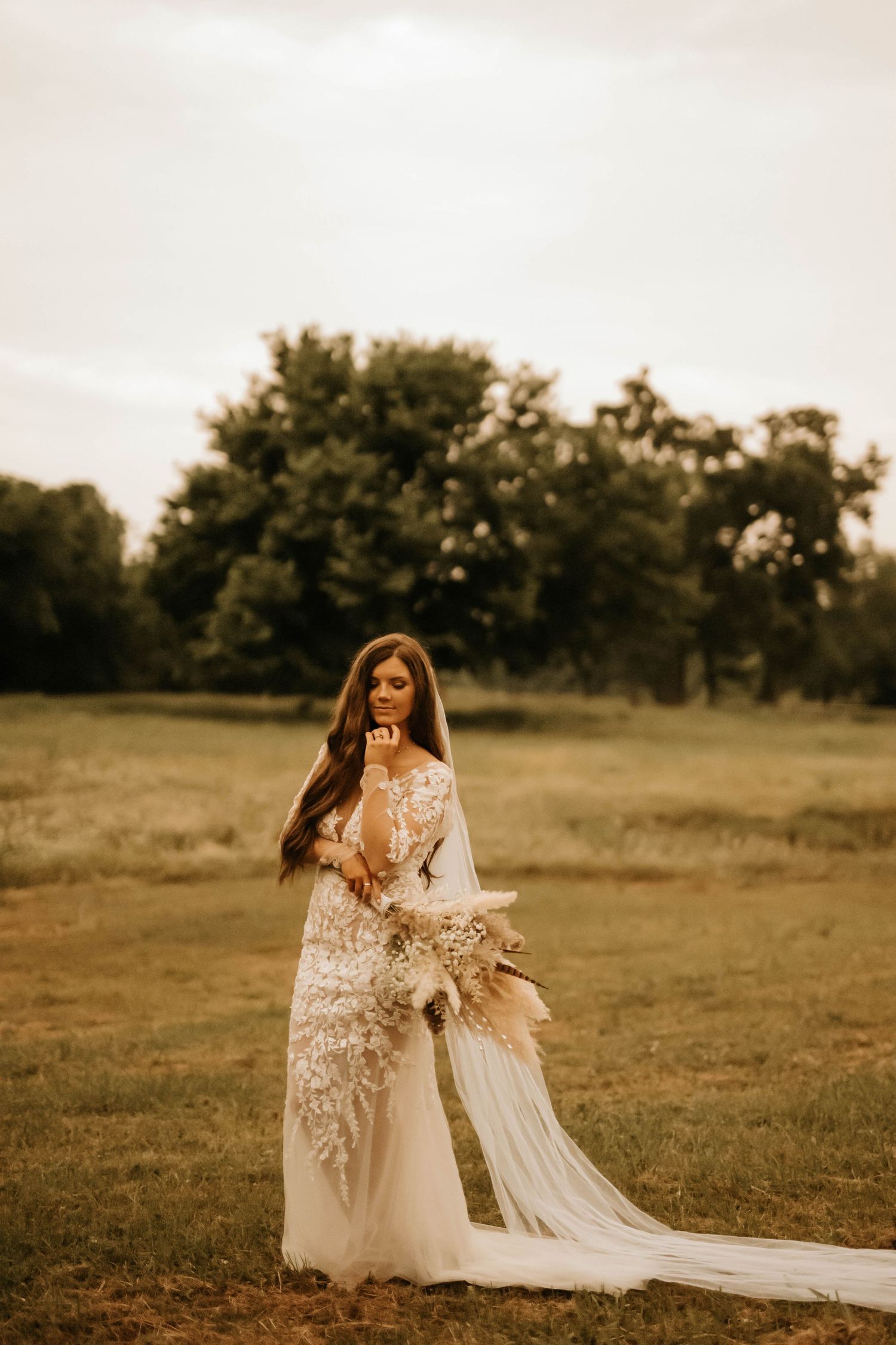 Bride in white lace gown with flowing veil poses in rustic field during golden hour.