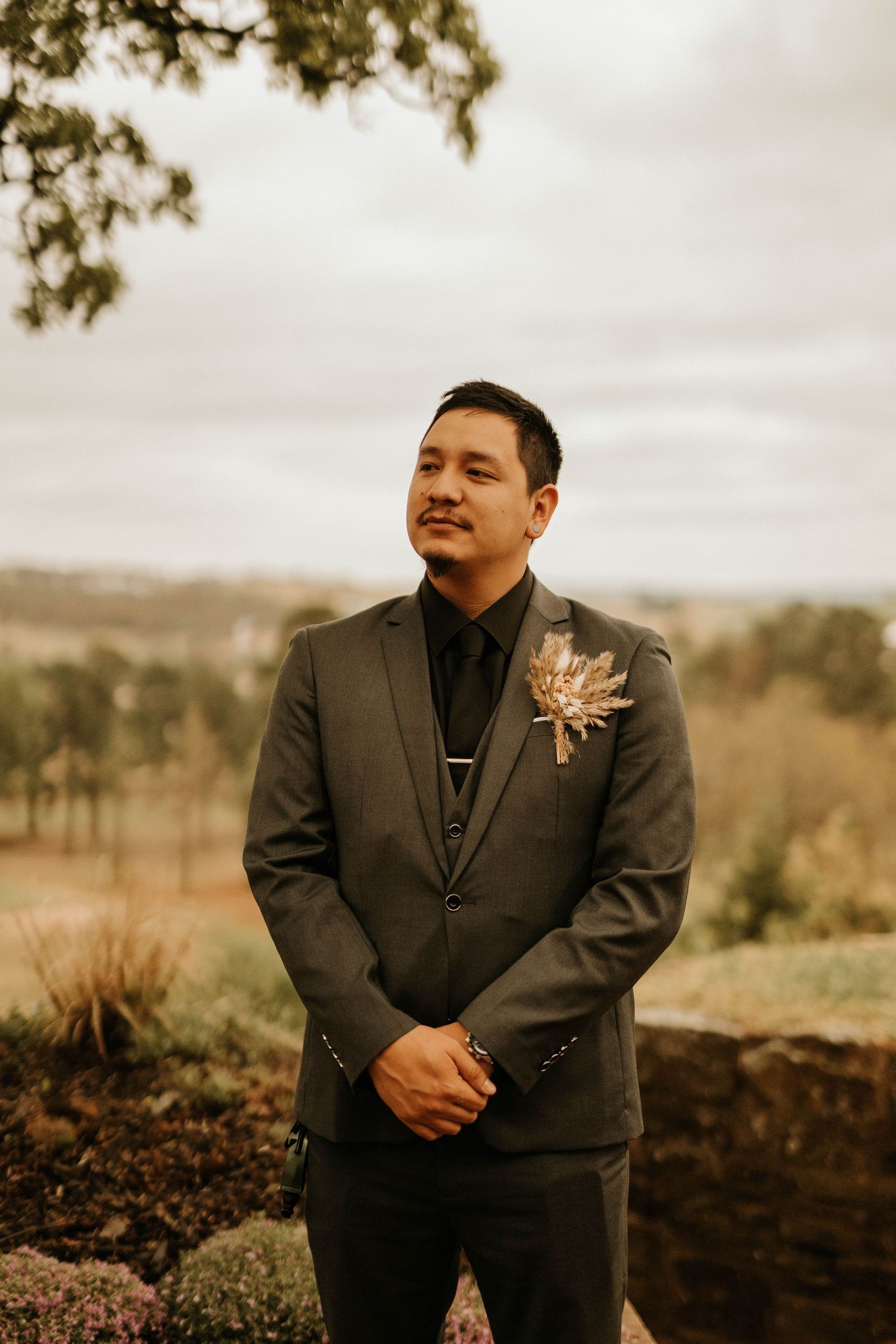 Groom in dark suit with boutonniere smiles against scenic autumn landscape.