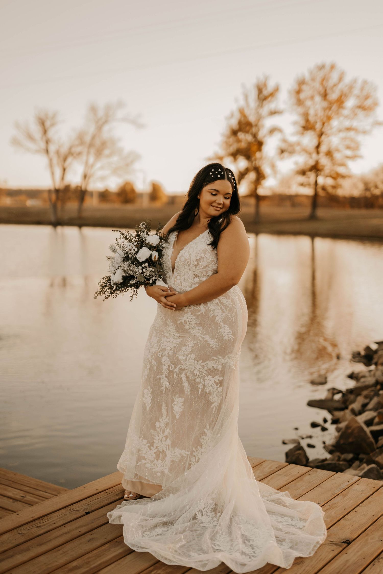 A bride in an elegant lace wedding dress holds a bouquet while standing on a wooden dock at sunset.