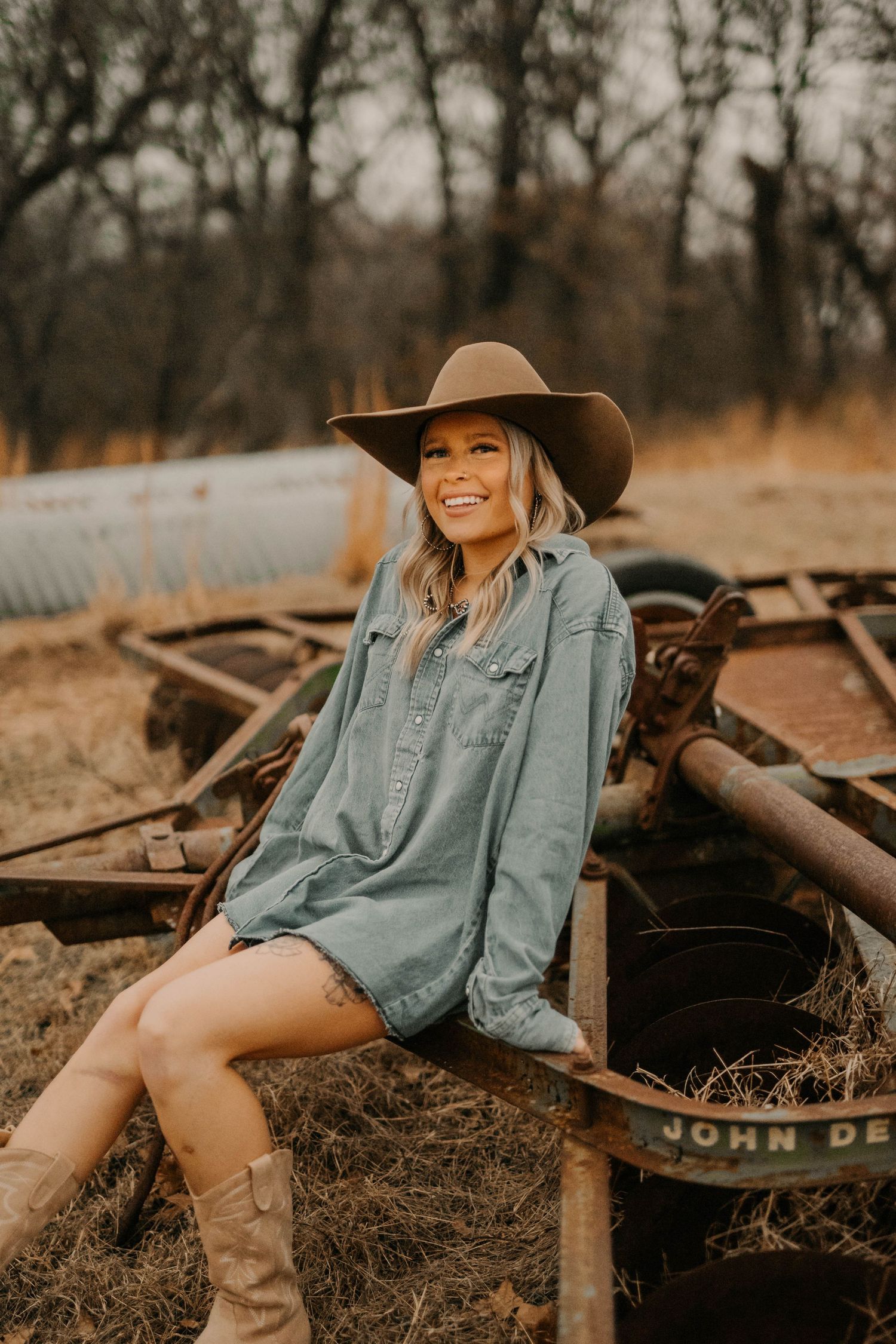 Person in denim shirt and cowboy hat sits on rustic farm equipment during golden hour.