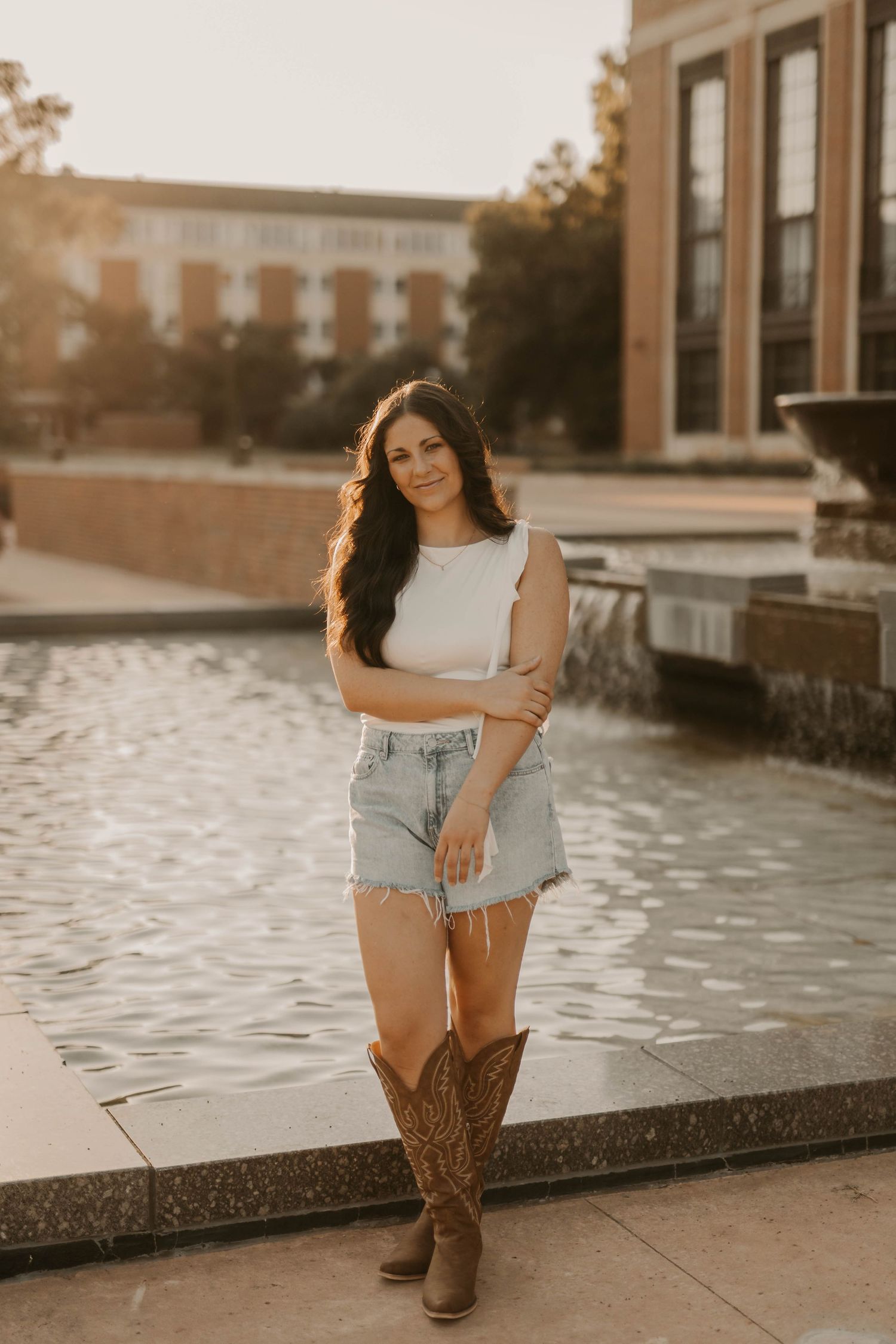 A person in cowboy boots and denim shorts stands by a fountain during golden hour at a university campus.