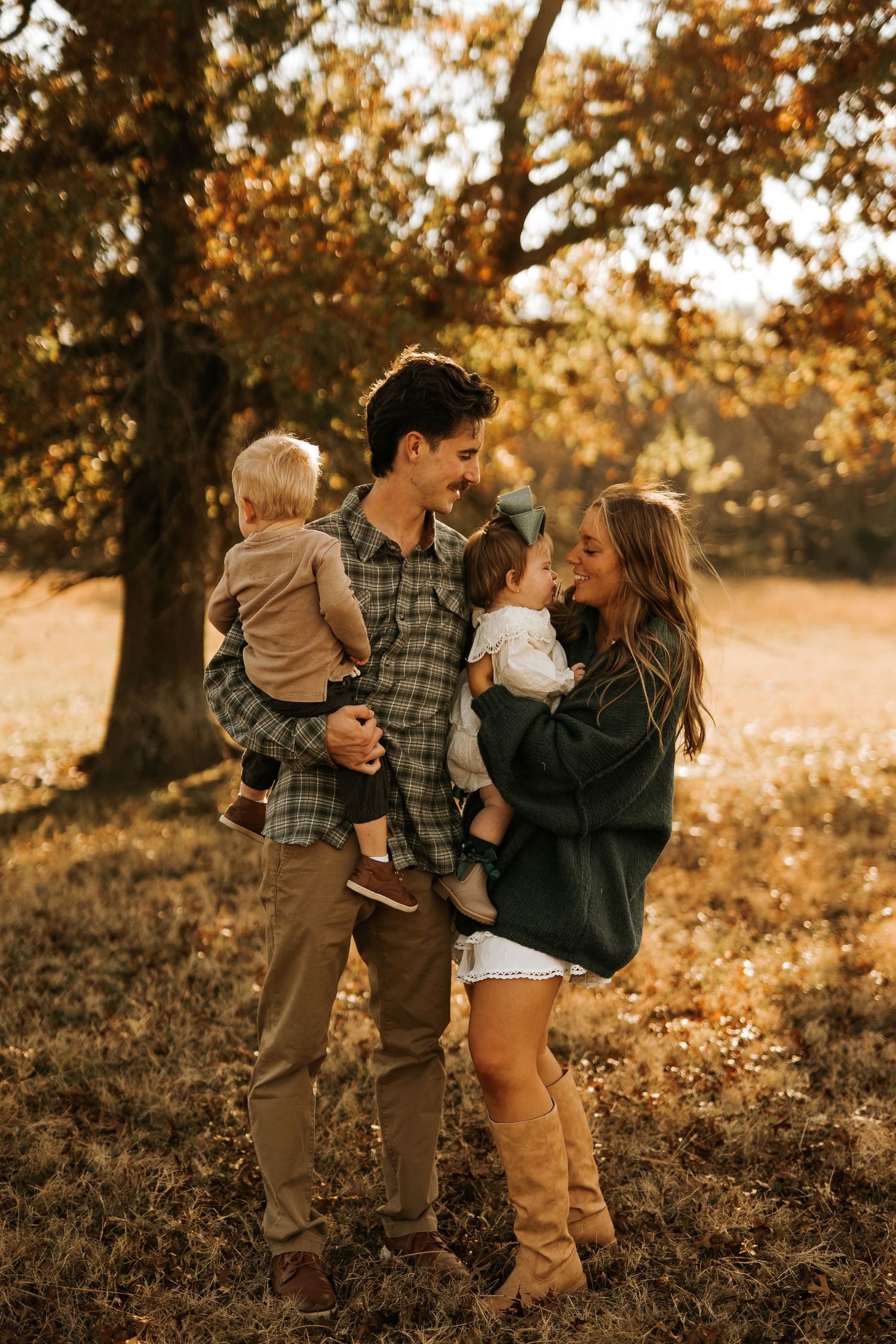 A young family of four poses outdoors in autumn, parents holding two toddlers, smiling warmly among golden fall foliage.
