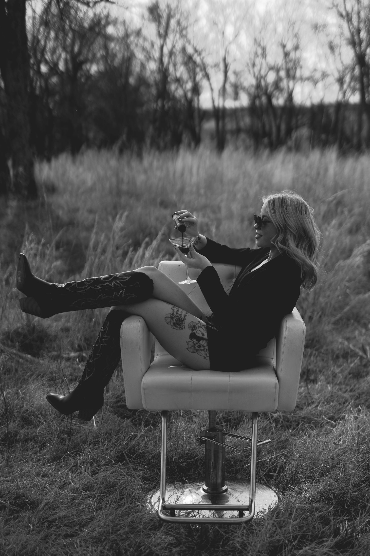 Black and white photo of a blonde woman in sunglasses sitting in a salon chair outdoors, holding a wine glass.
