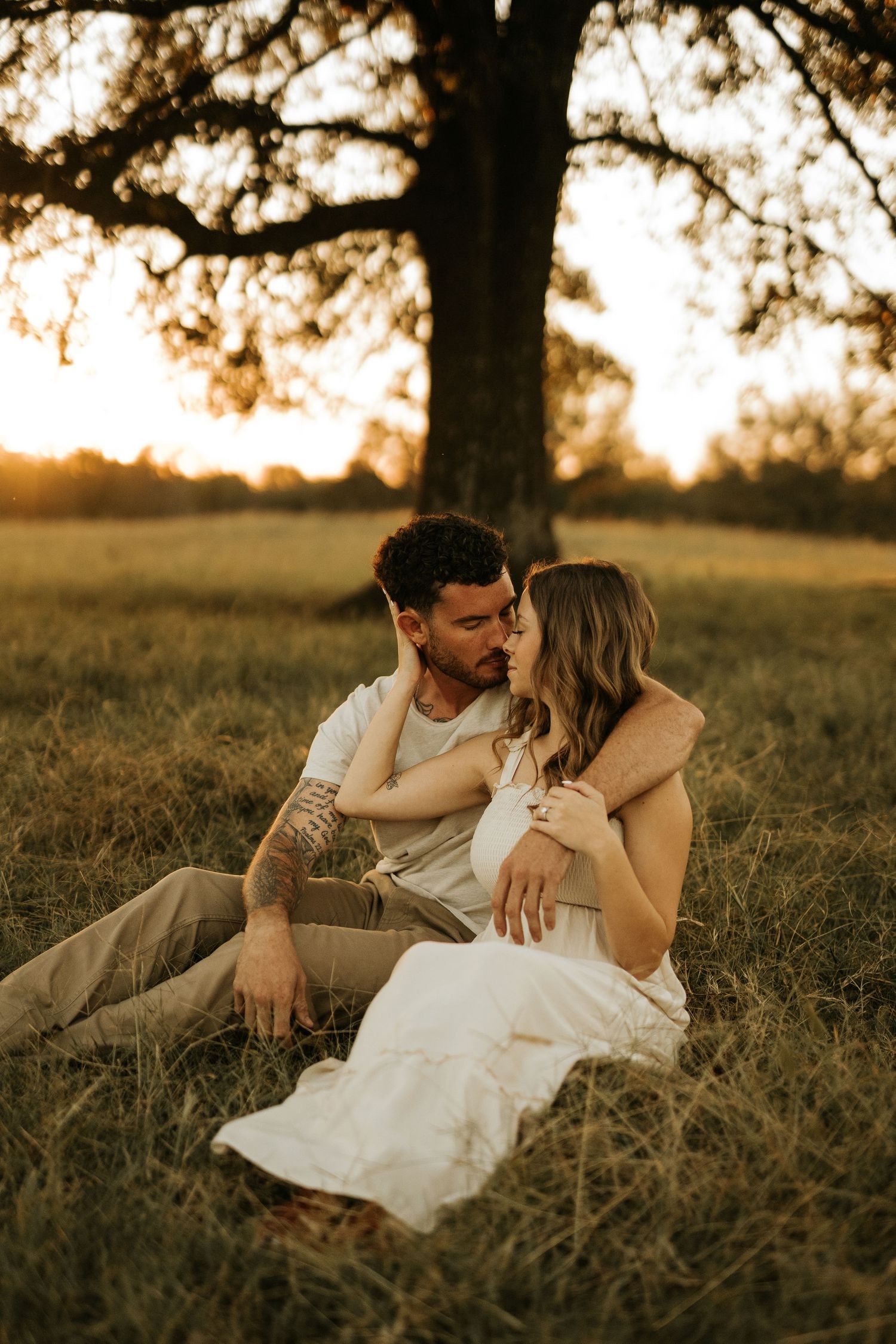 Romantic couple sitting together in golden sunset light beneath a large oak tree in a meadow.