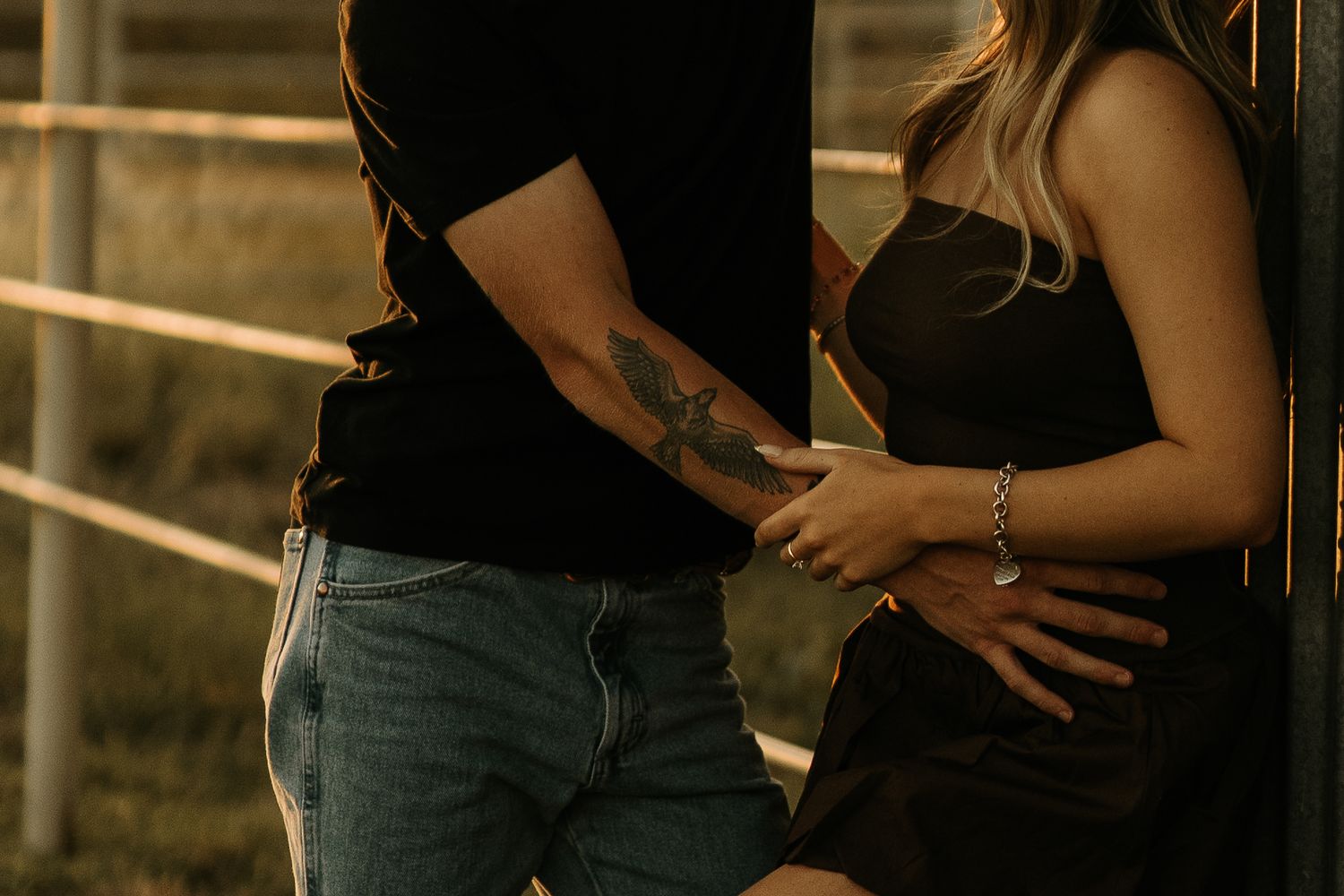 Couple standing close together, man with tattoo on arm wearing jeans, woman in black dress, romantic outdoor setting.