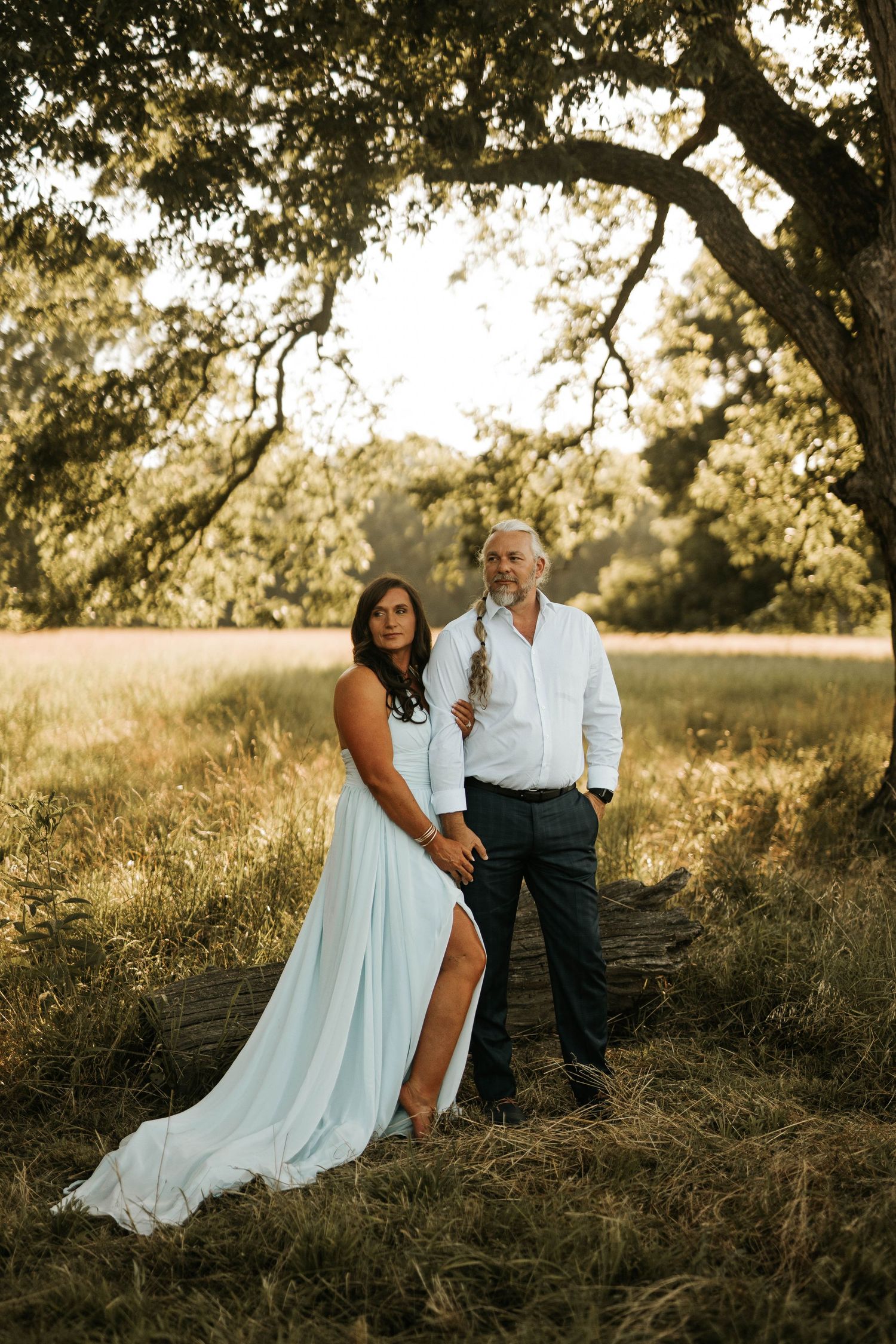 Couple poses under oak tree in golden field, woman in light blue gown, man in white shirt, sunset backdrop.