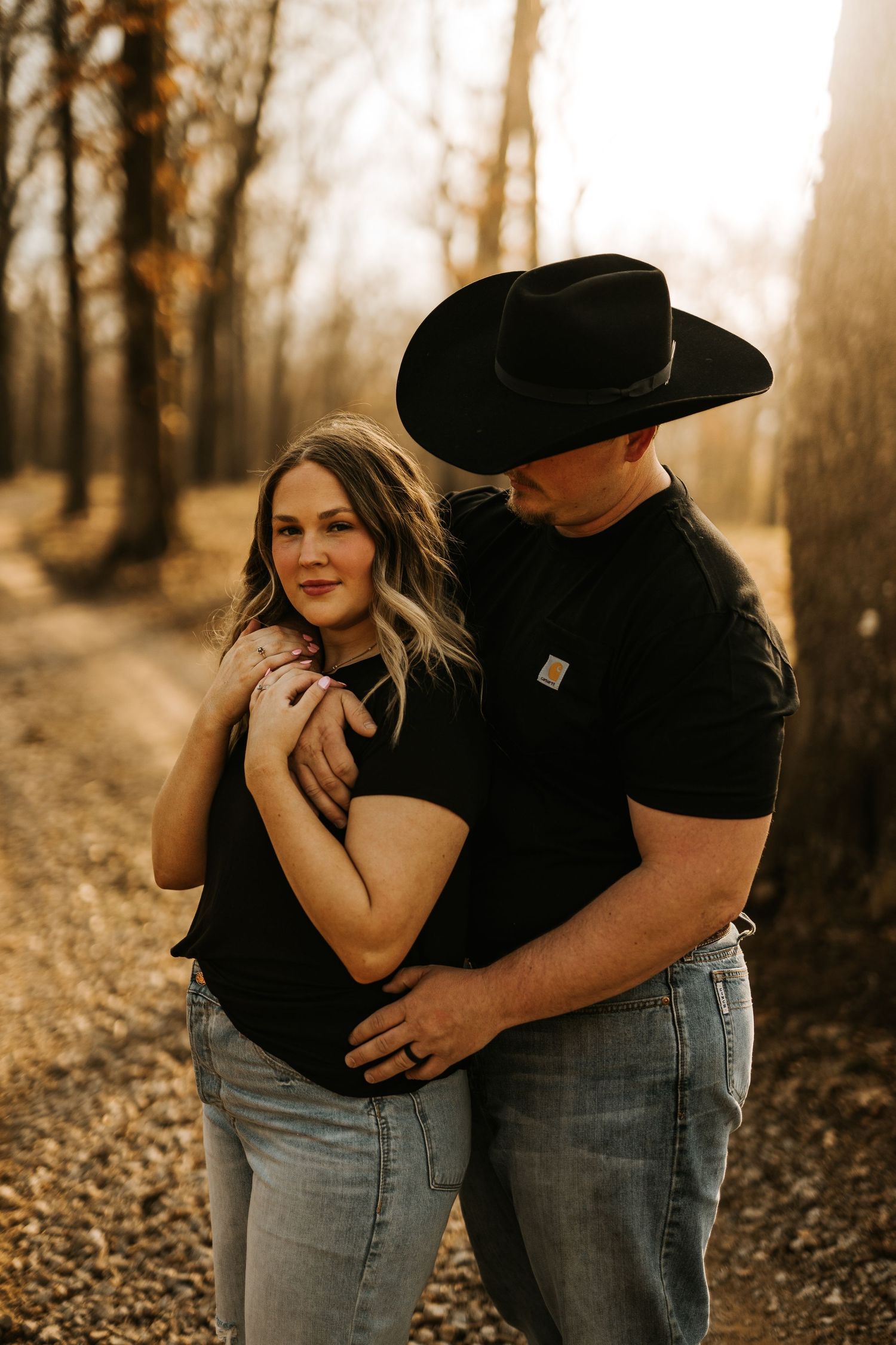 A couple embraces on a sunlit forest path, man in black Carhartt shirt and cowboy hat, woman in black top and jeans.
