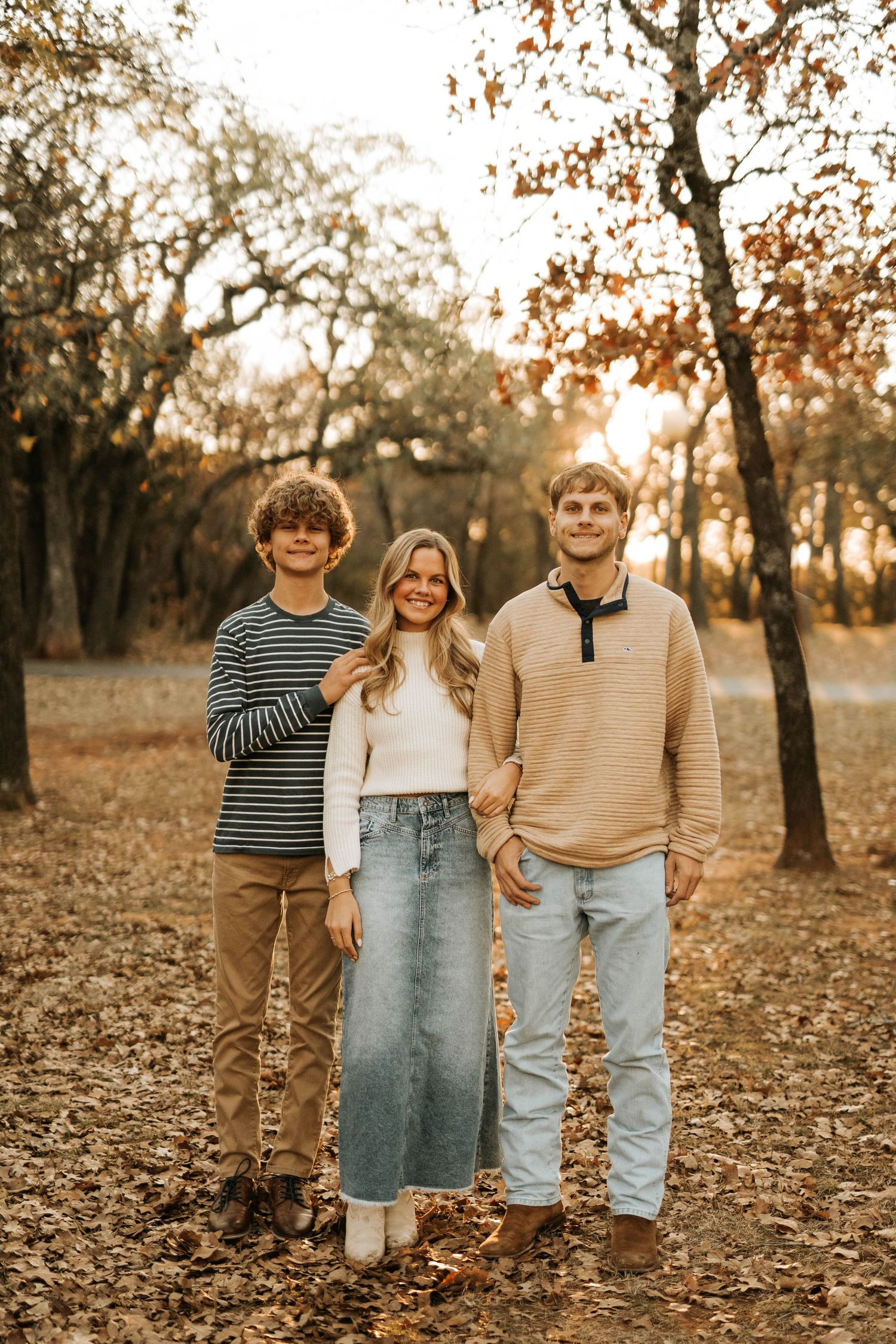 Friends pose together on fallen leaves in autumnal forest setting during golden hour.