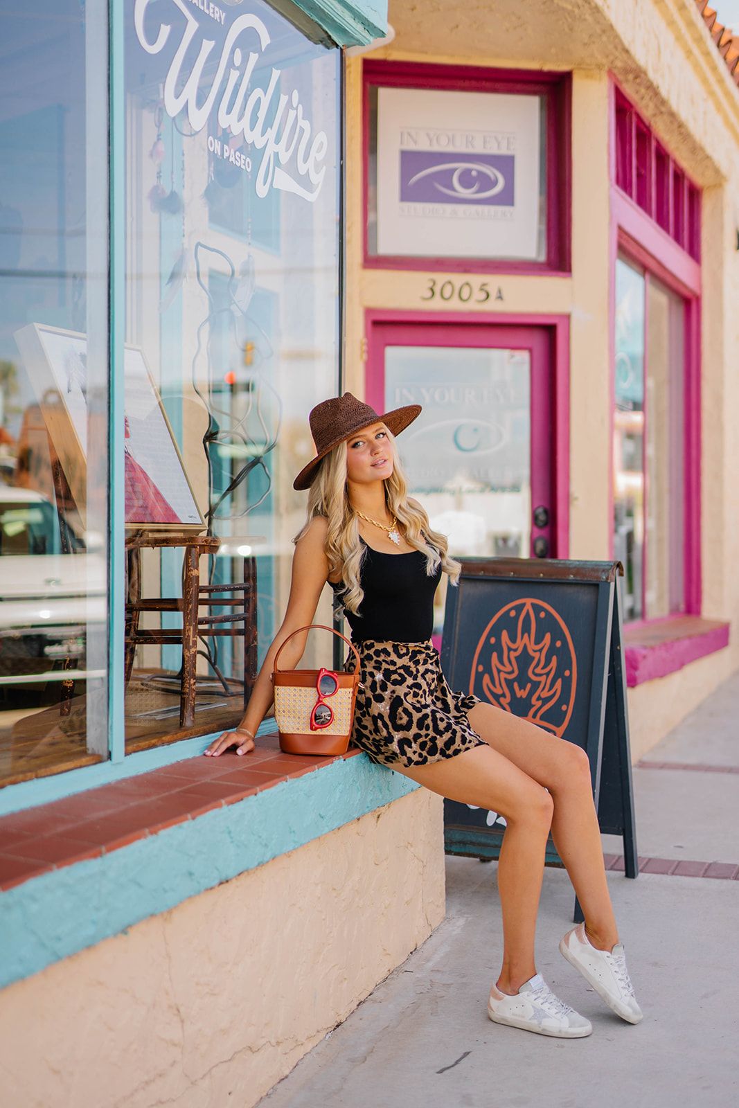 Woman in black top and leopard skirt sits casually on storefront ledge with vintage signage.