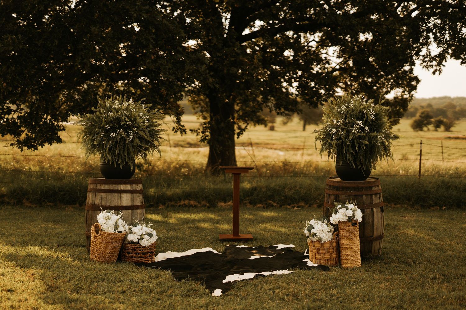 Rustic outdoor wedding ceremony setup with barrel stands, potted greenery, wooden cross, and cow hide rug on grass.