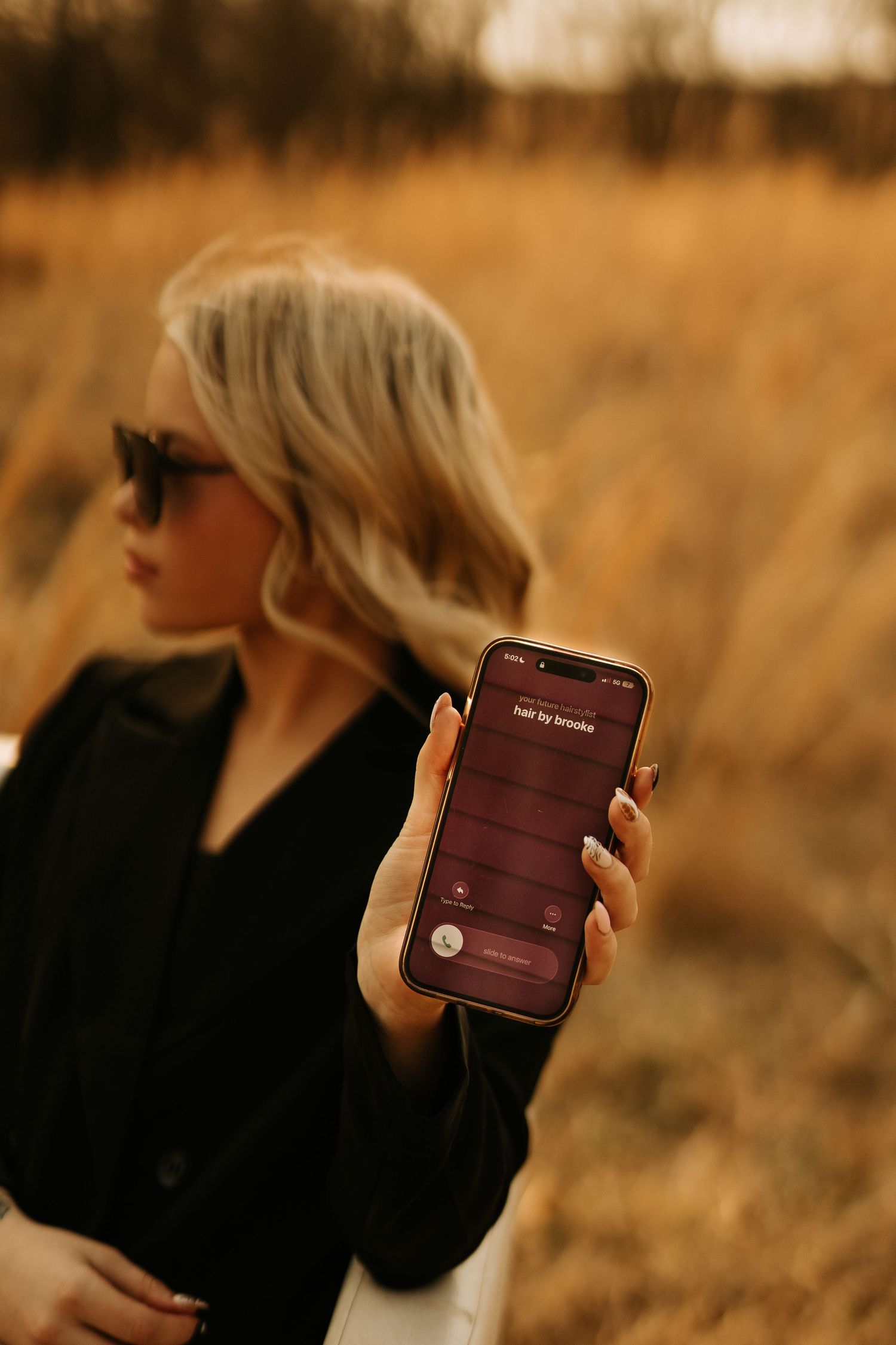 Blonde woman in black jacket holding smartphone displaying a dark red app screen in a golden field.