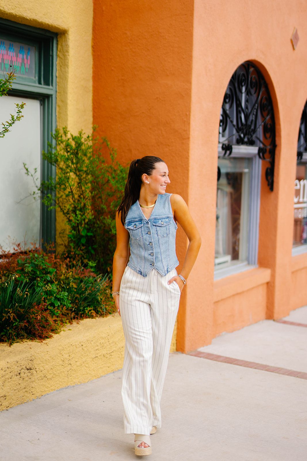 Casual summer outfit featuring denim vest and white pants against peach colored building.