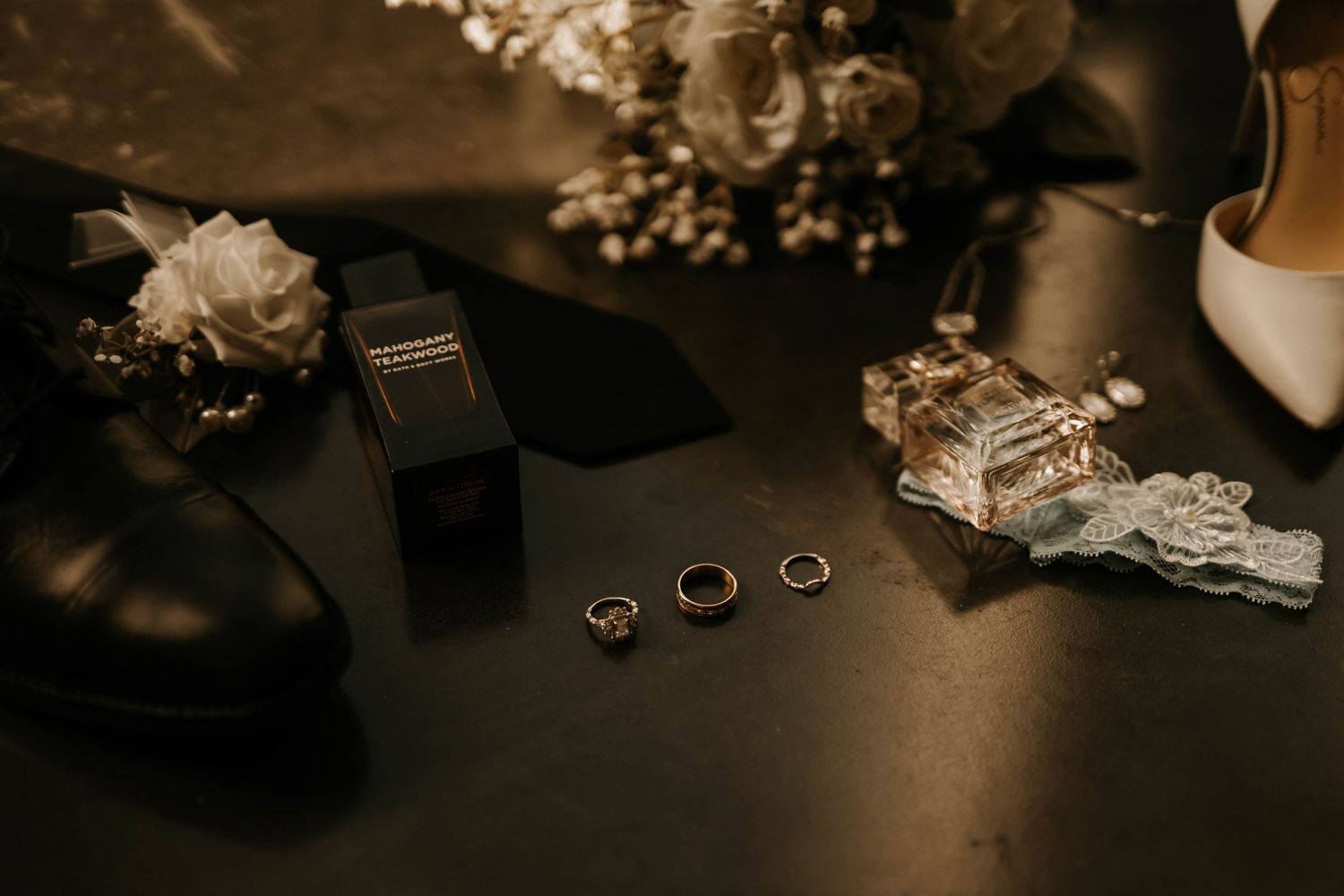 Wedding details including rings, shoes, and delicate floral arrangements artfully arranged on a dark surface.