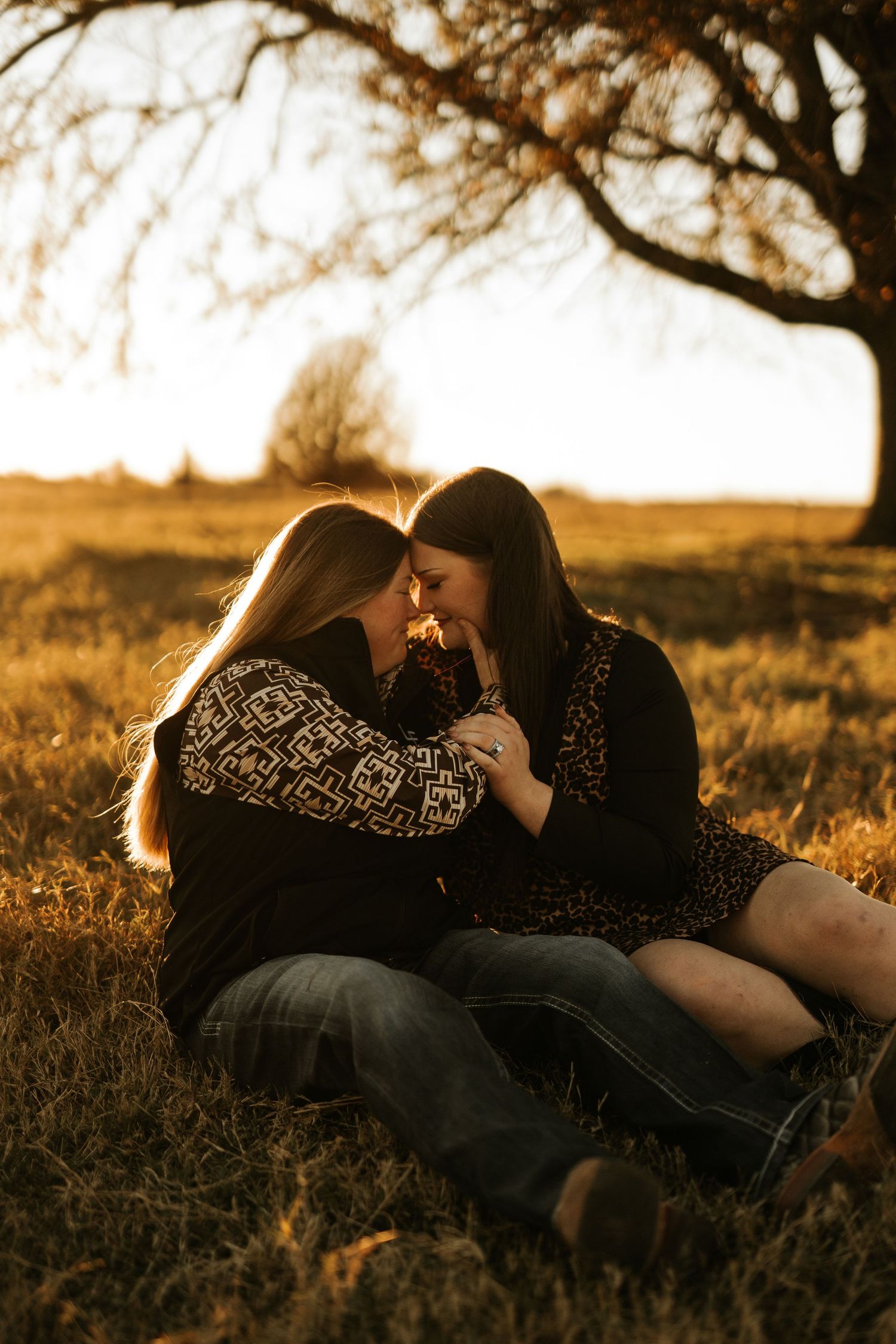 Two women sitting in golden field at sunset, foreheads touching in a romantic embrace near a bare tree.