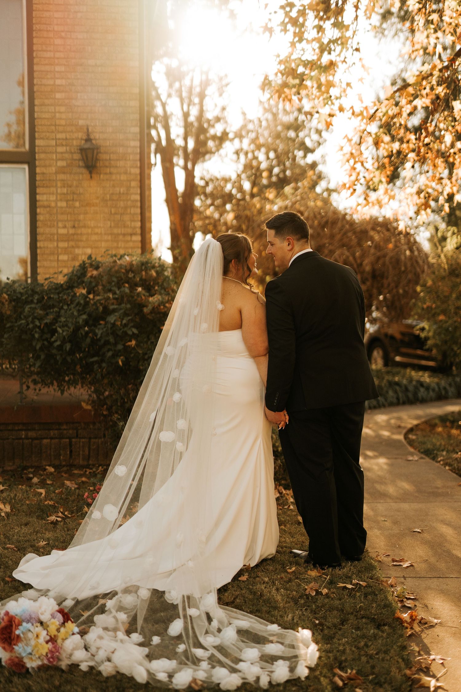 Bride in white gown with floral veil and groom in black tuxedo share a romantic moment outdoors in golden autumn light.