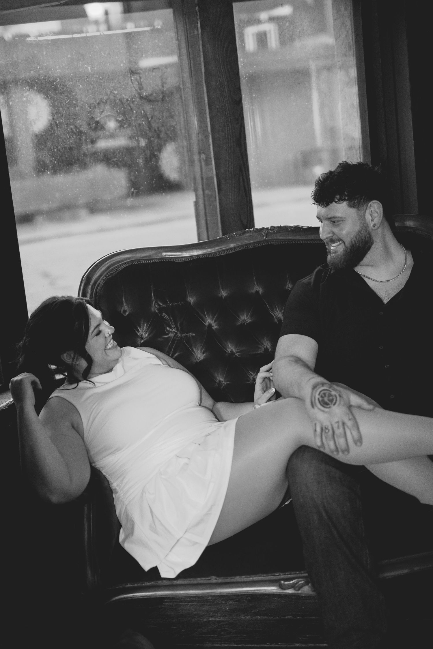 Black and white maternity photo of couple on vintage sofa, man touching pregnant woman's belly, intimate moment.
