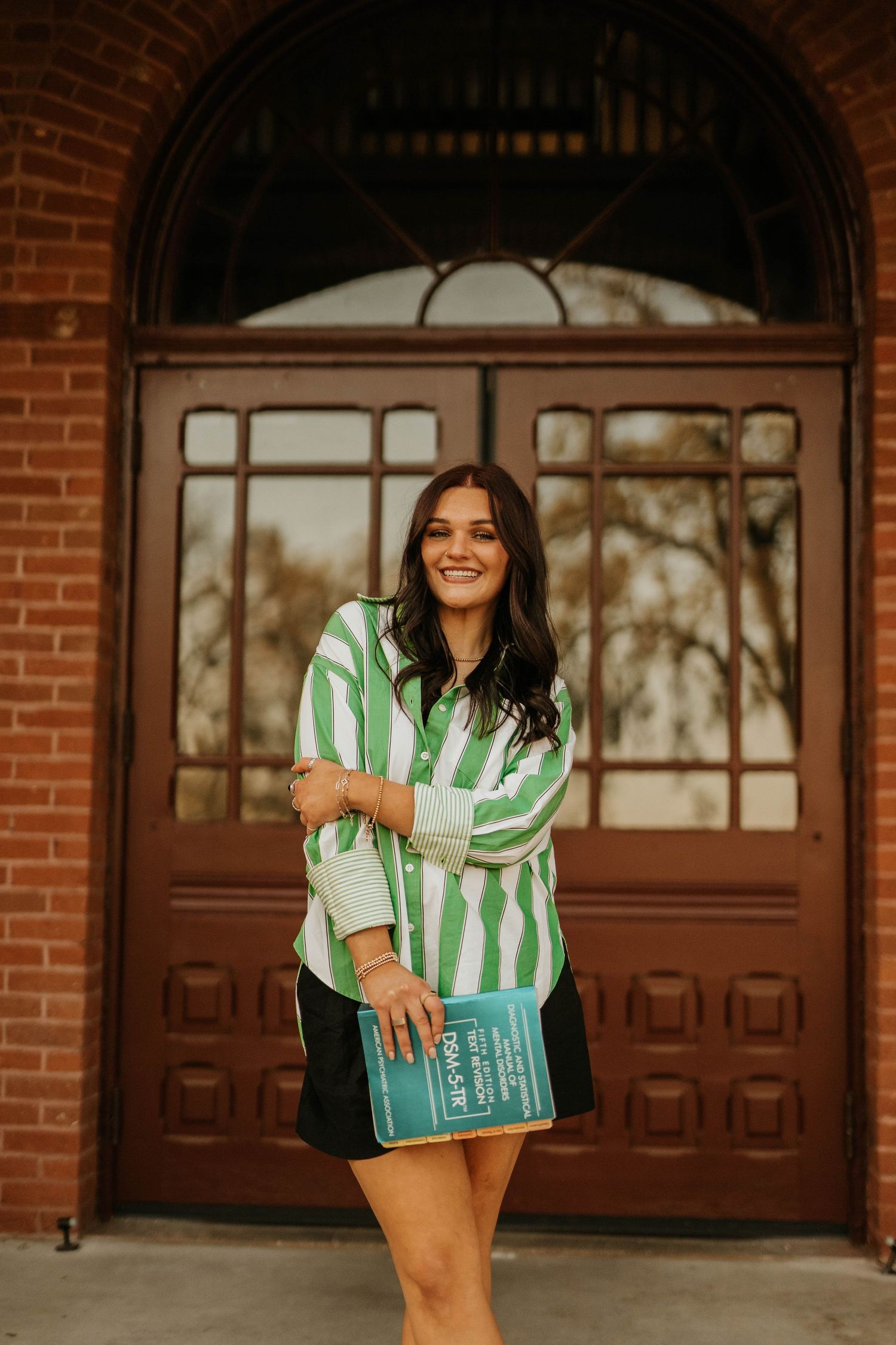 Graduate in green striped shirt poses with book in front of ornate wooden doors.