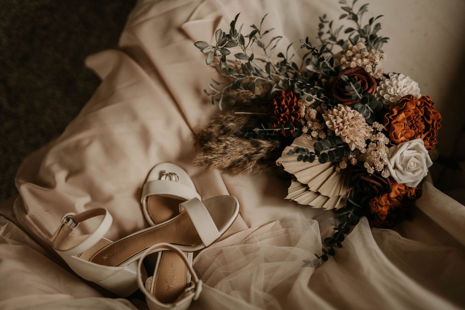 Wedding detail shot featuring bridal shoes and a dried flower bouquet on cream-colored fabric.