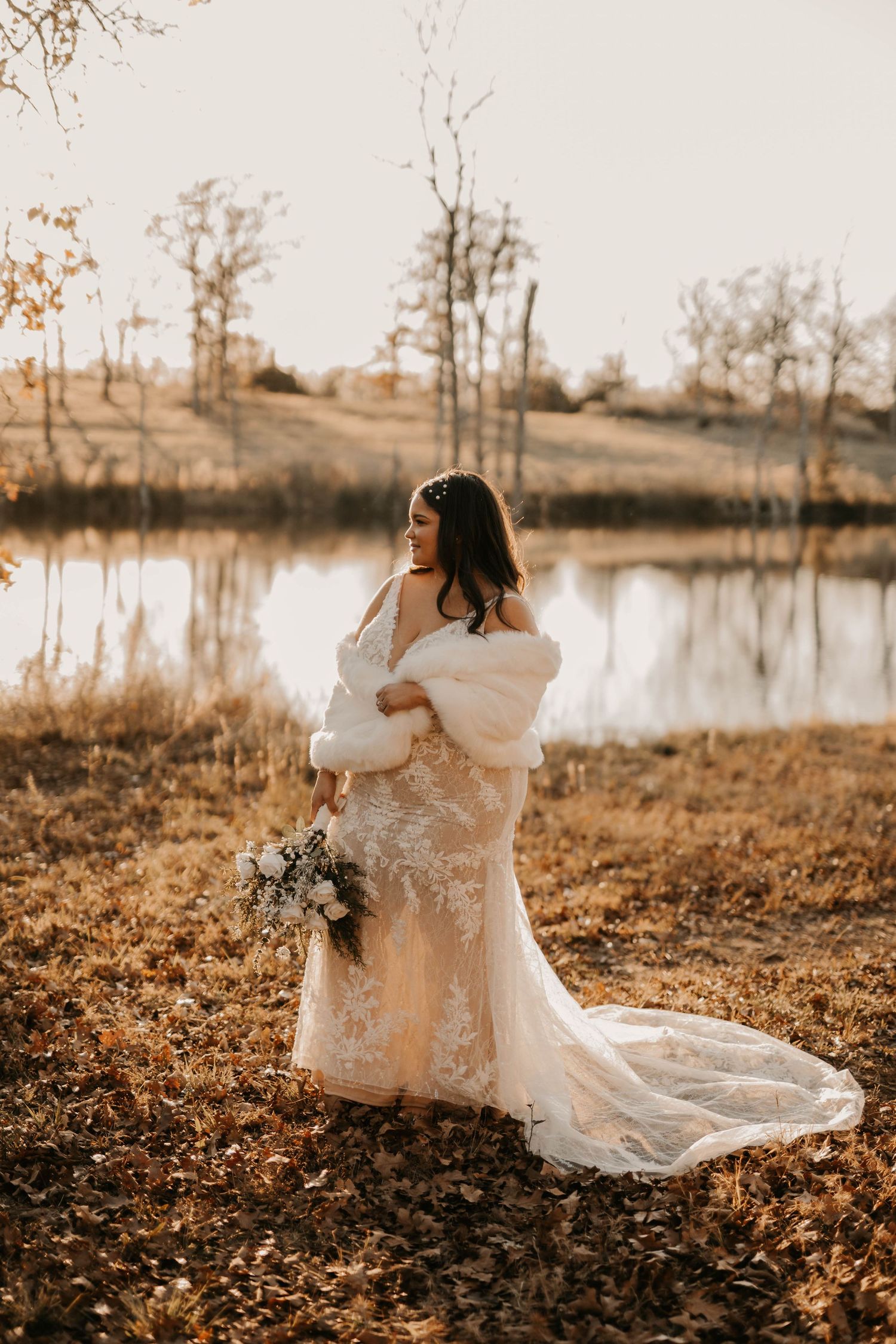 A bride in a lace gown and fur wrap poses near a tranquil lake during a golden sunset photoshoot.