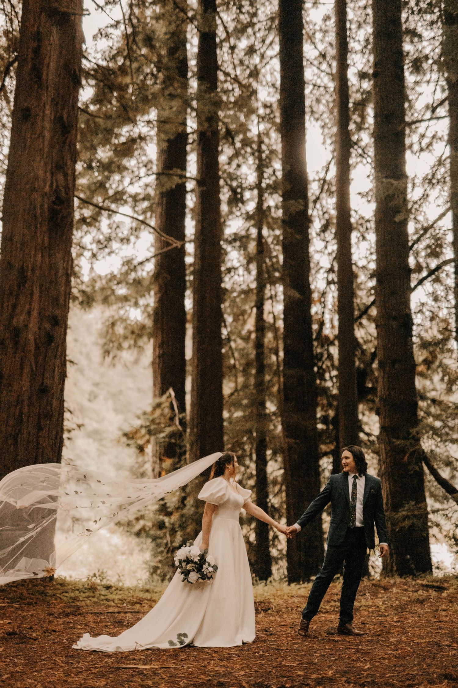 A couple in wedding attire poses dramatically among tall redwood trees.