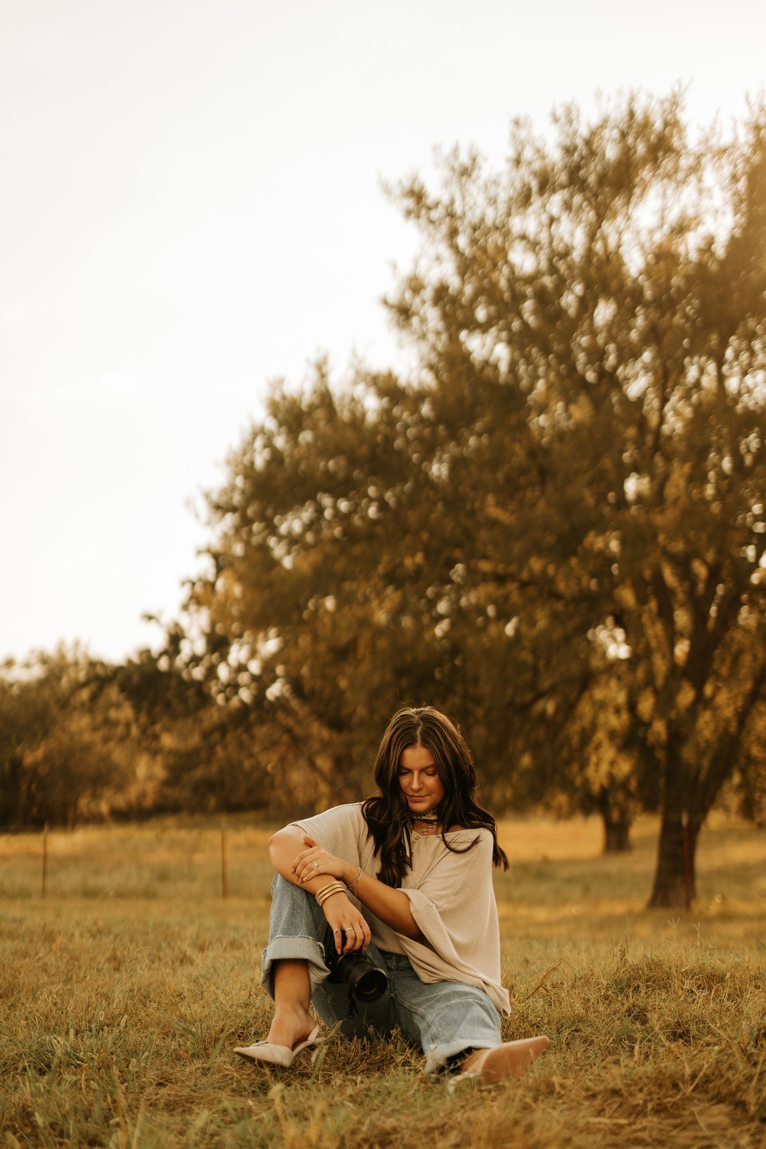 Woman sitting in golden field at sunset, holding camera, with trees in warm backlight, wearing jeans and white shirt.