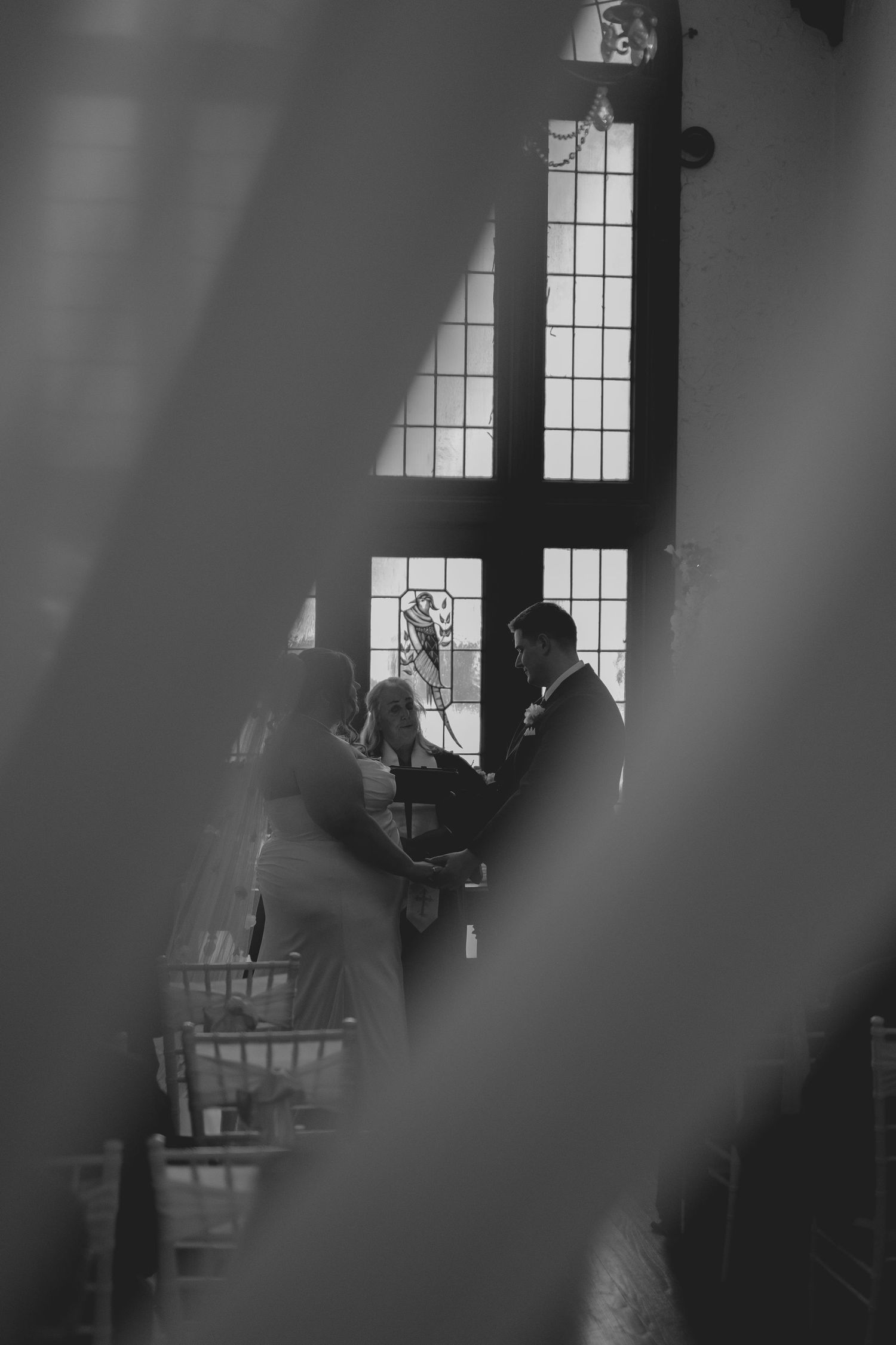 Black and white wedding ceremony photo through floral foreground, couple exchanging vows before stained glass windows.