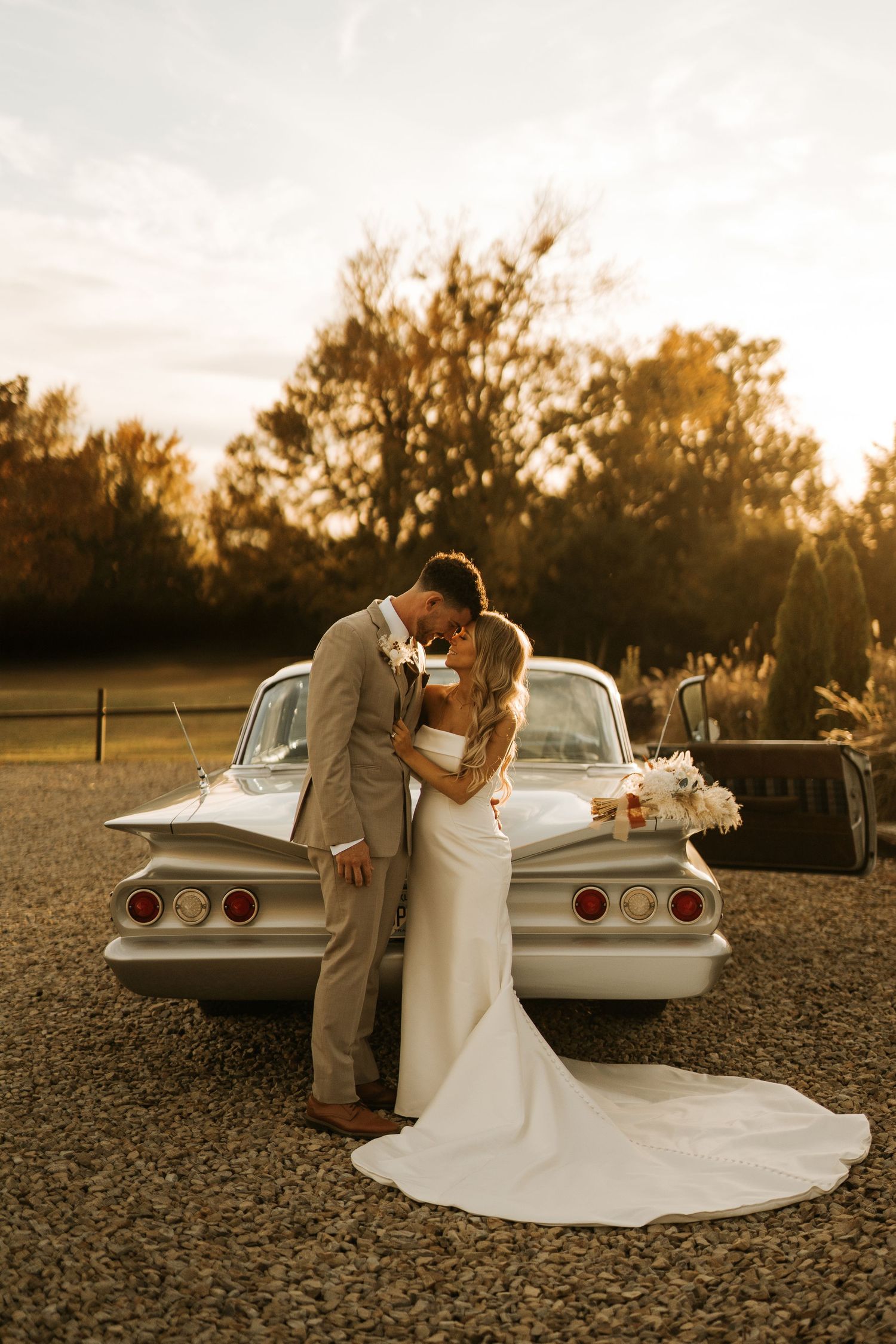 A bride and groom share a romantic moment leaning in for a kiss beside a vintage silver car at golden hour.