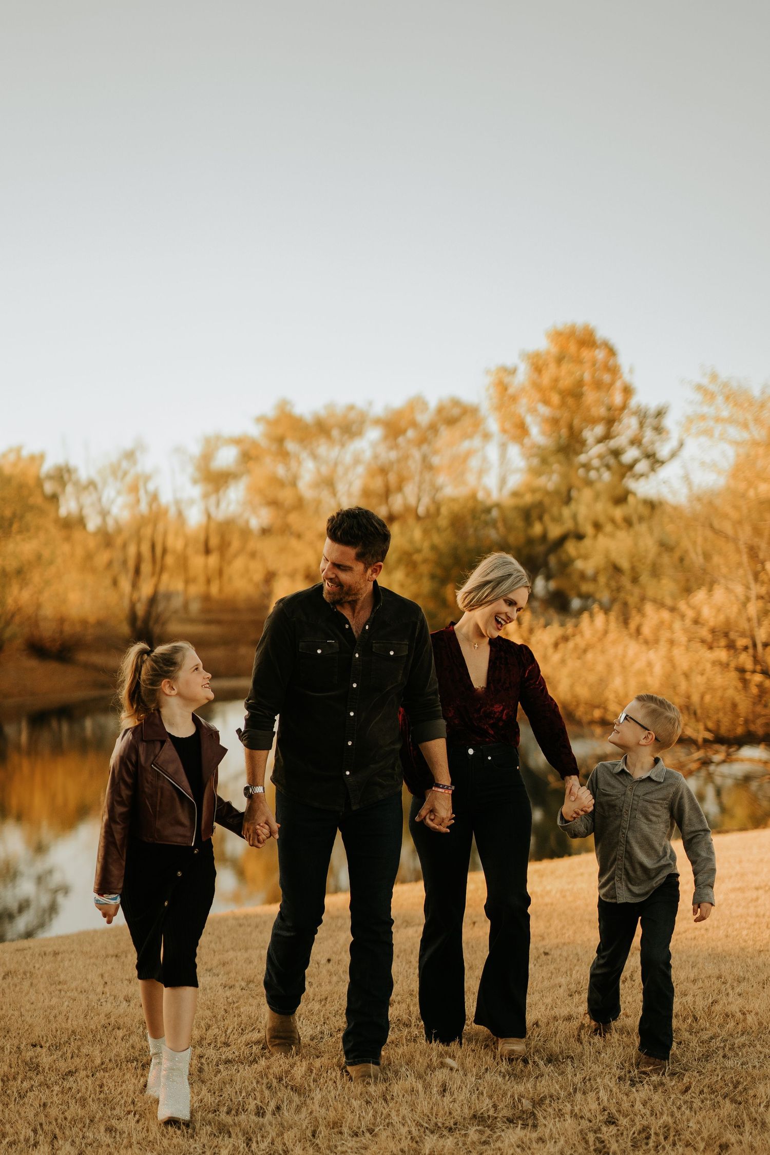 Family of four holding hands walking through a golden autumn field near a pond at sunset.
