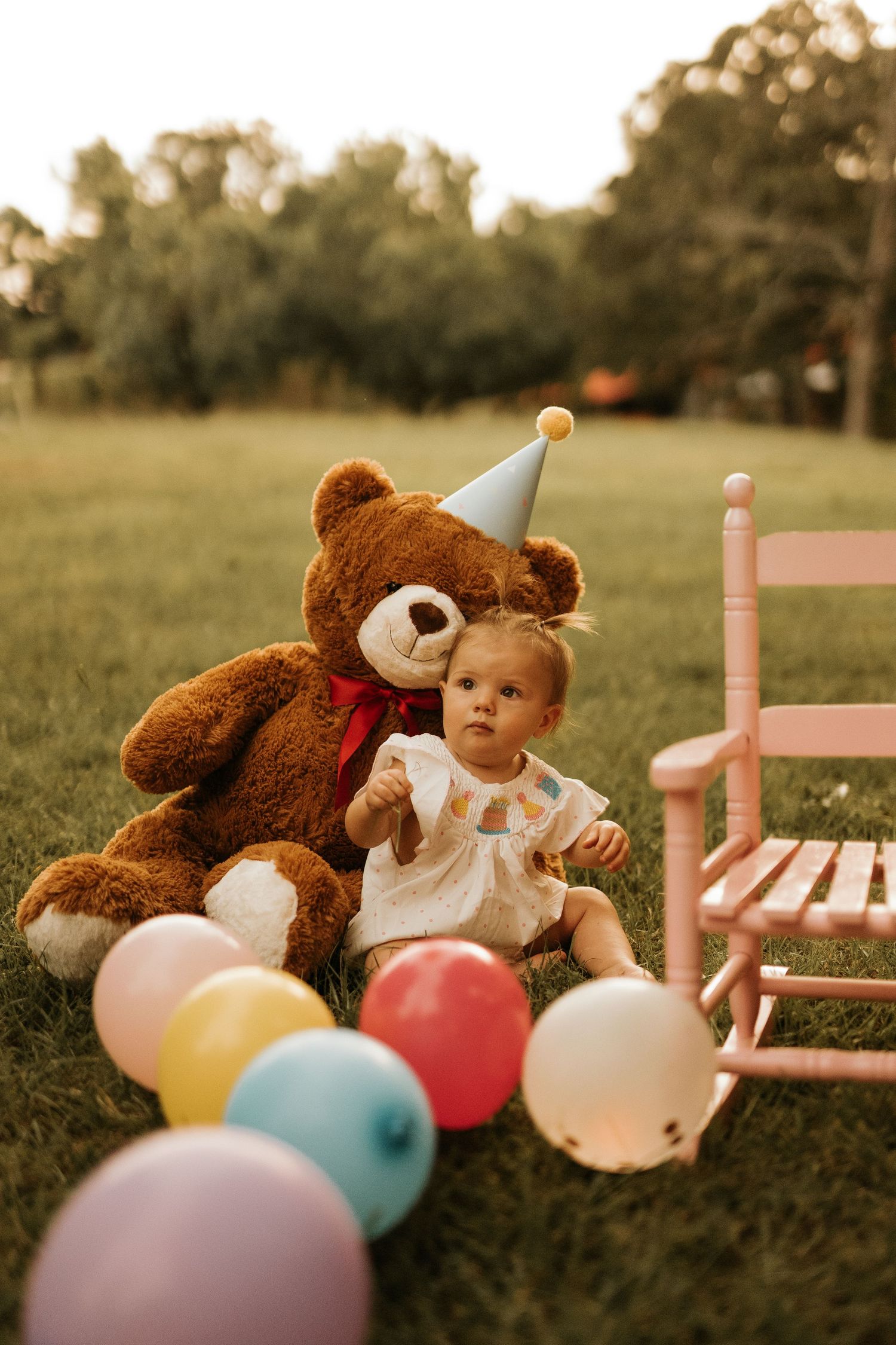 Baby girl sitting on grass with a large teddy bear wearing a party hat, colorful balloons, and a pink rocking chair.