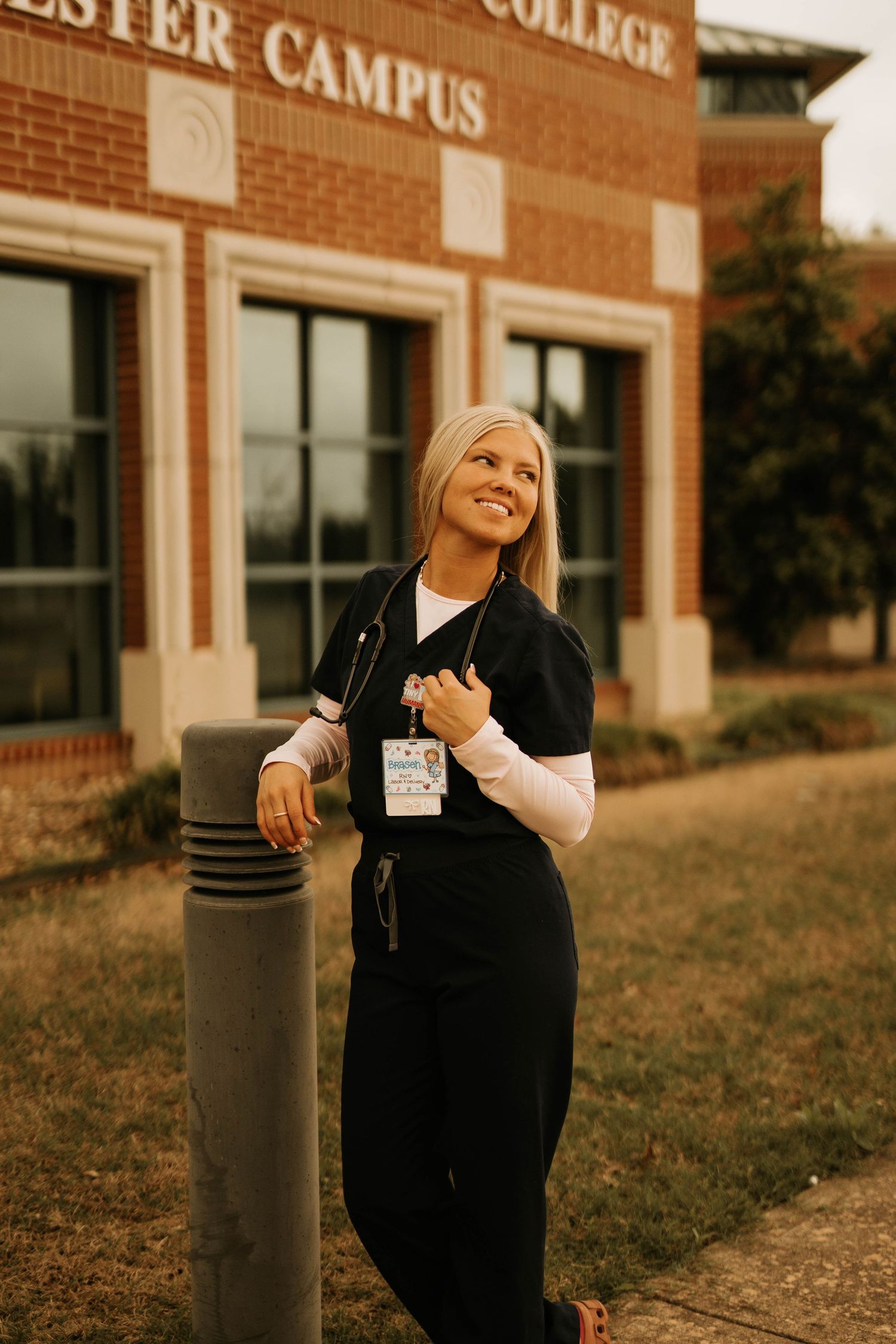 Someone in medical scrubs stands smiling outside a college campus building.