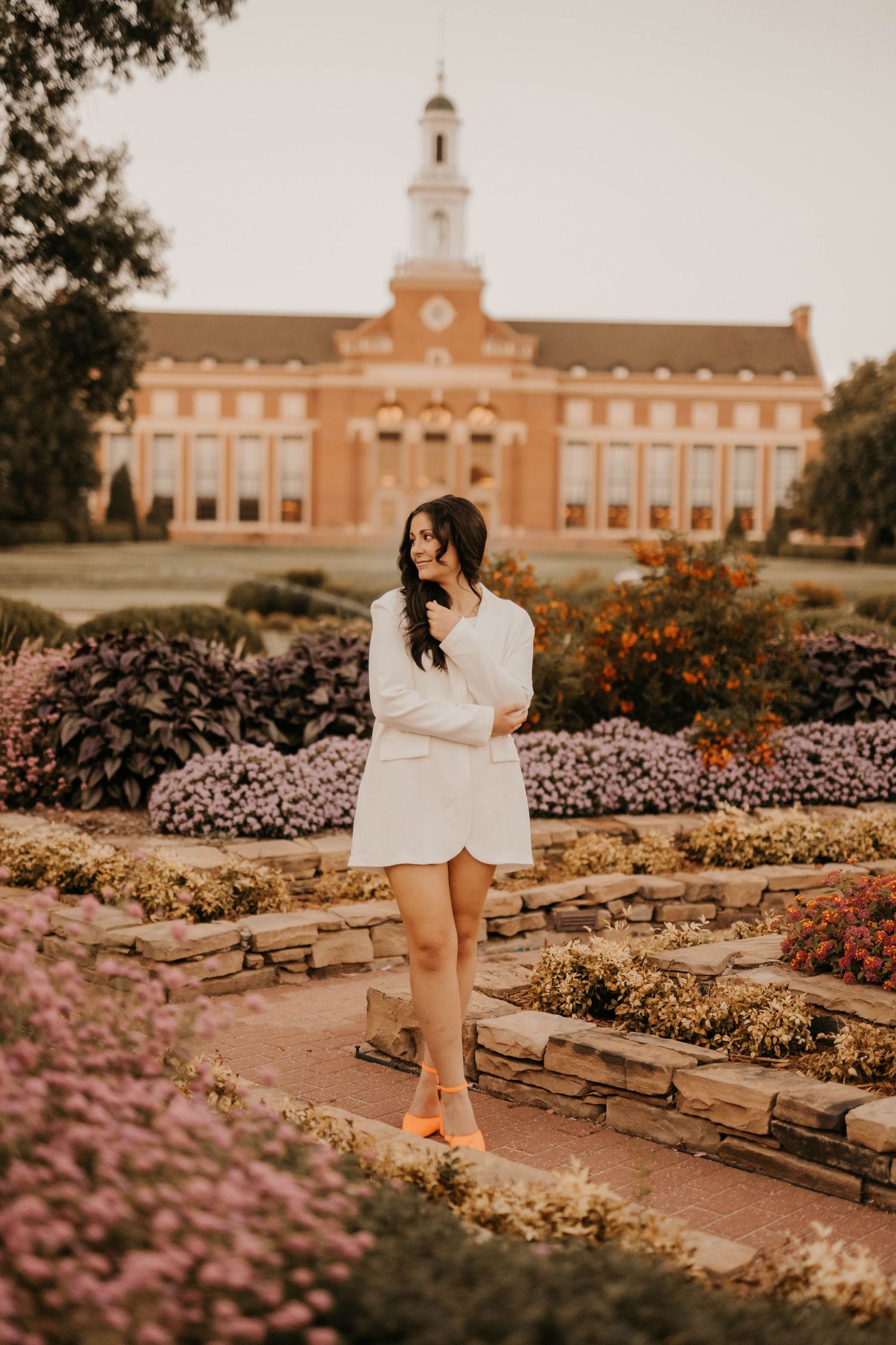 A person poses in a white outfit among flower gardens with a grand collegiate building in the background.